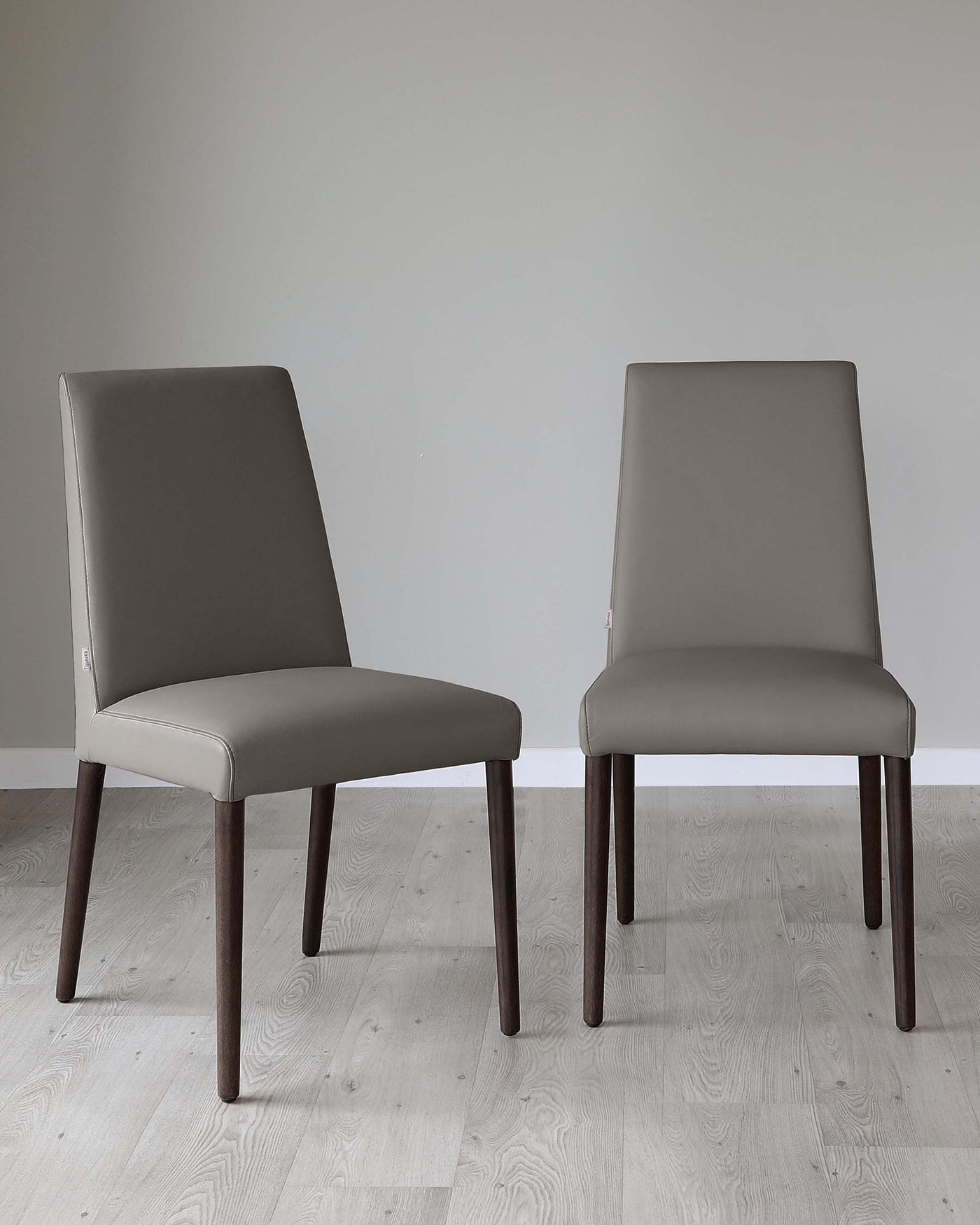 Two modern dining chairs with sleek, taupe upholstery and dark wooden legs, standing on a light wood floor against a soft grey background.
