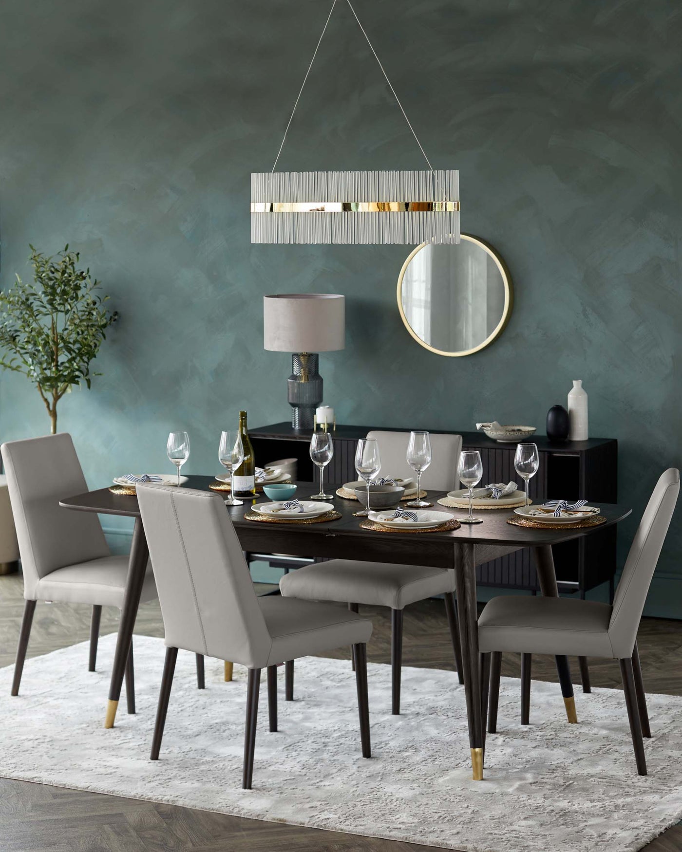 Contemporary dining set featuring a rectangular table with dark wooden finish and tapered legs tipped with brass caps, surrounded by six upholstered chairs in a matching grey fabric. The table is set with elegant dinnerware, glasses, and a bottle of wine, ready for a sophisticated meal. A large rectangular pendant light with vertical slats and golden accents hangs above, complementing the modern, stylish aesthetic of the dining area.