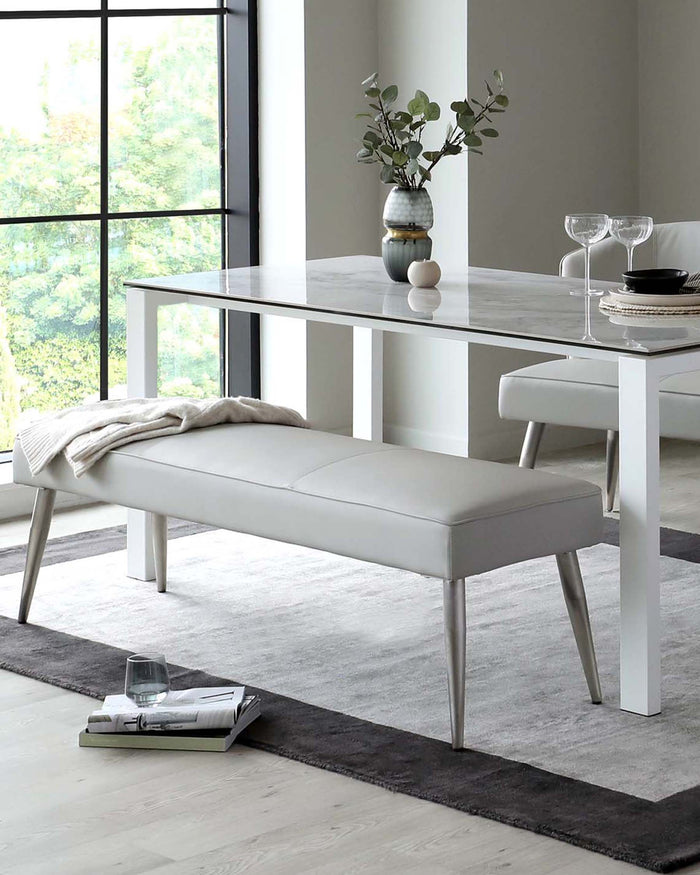 Modern minimalist dining room furniture featuring a sleek white marble top table with metal legs and a matching upholstered bench in light grey, set on a textured grey area rug.