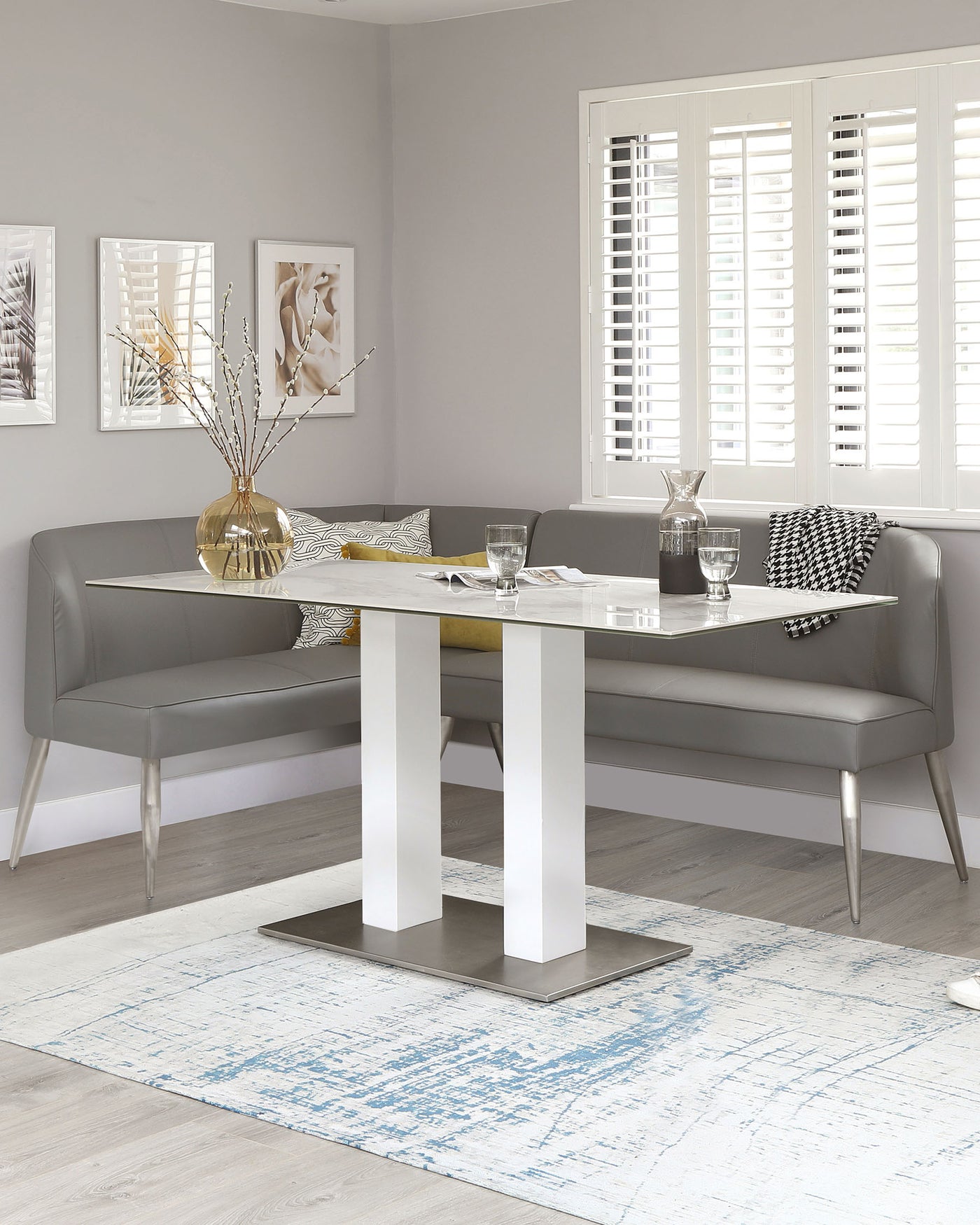 Modern dining area featuring a rectangular table with a glossy white finish and metallic silver legs, paired with a long, grey upholstered bench with sleek lines and silver metallic legs. The setting is accented with contemporary home decor and situated on a distressed blue and white area rug.