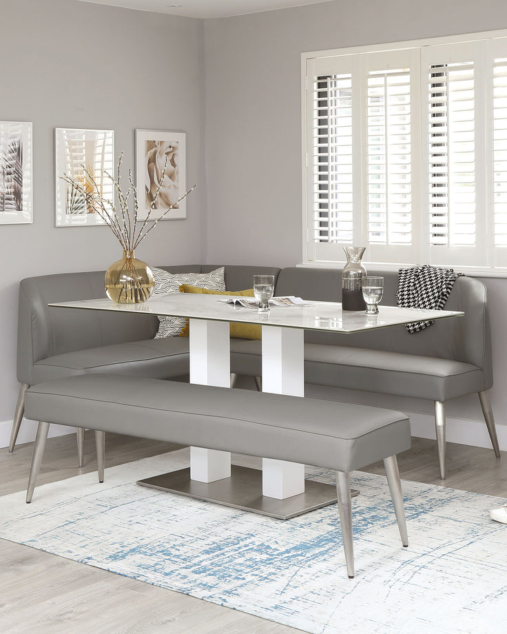 Modern minimalist dining set featuring a white rectangular table with sleek metallic accents and two matching grey upholstered benches with angled metal legs, arranged on a distressed blue and white area rug. The table is adorned with decorative items including a gold vase with branches, and the benches are accented with patterned throw pillows.