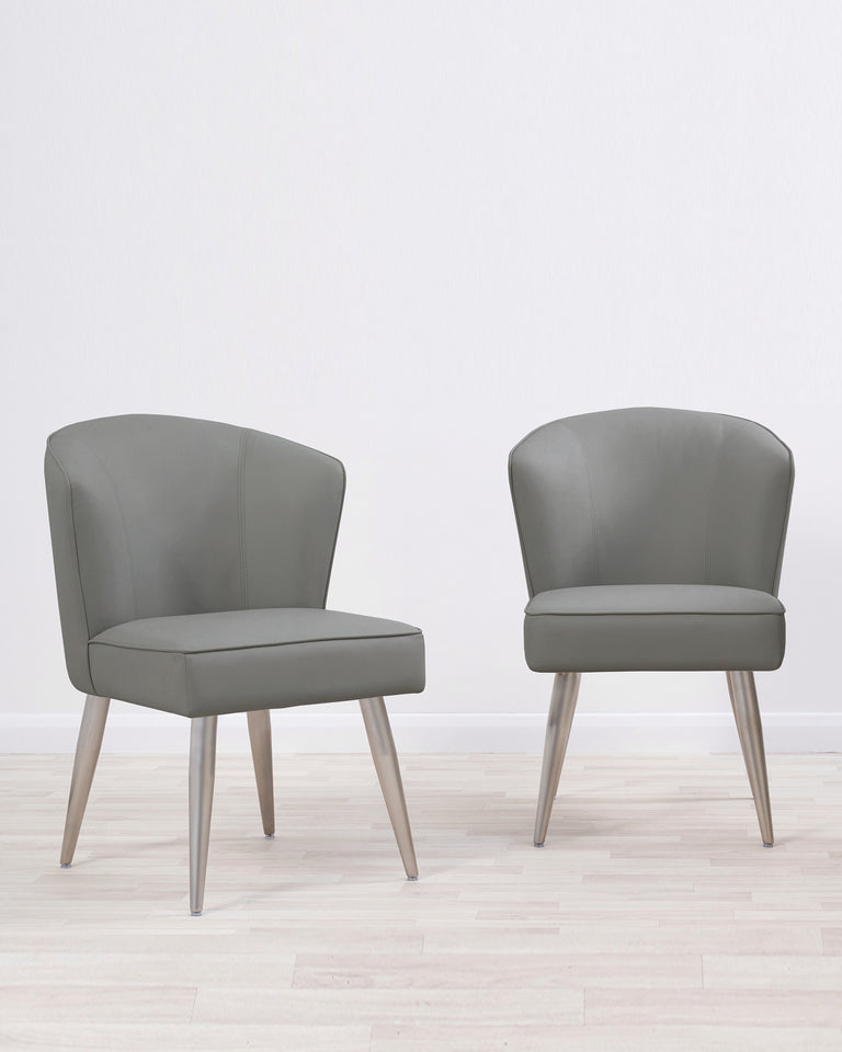 Pair of contemporary grey upholstered accent chairs with curved backrests and angled wooden legs on a light hardwood floor against a white backdrop.