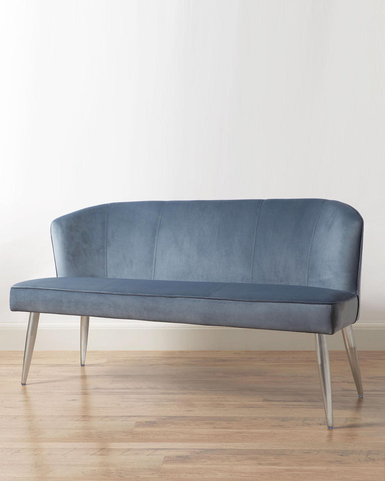 Elegant modern blue velvet settee with a gently curved backrest and sleek silver metal legs, placed against a neutral wall on a hardwood floor.
