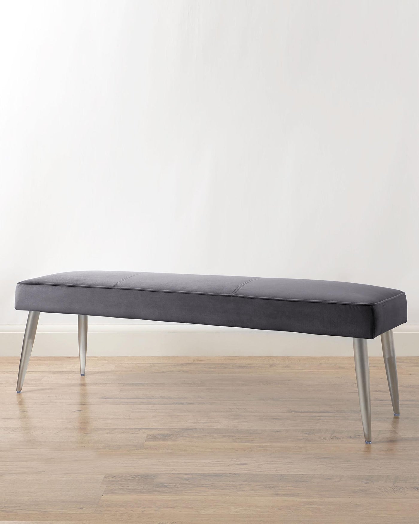 Modern upholstered bench with sleek, charcoal grey fabric and slender, tapered metal legs in a brushed finish, positioned against a neutral wall and hardwood floor.