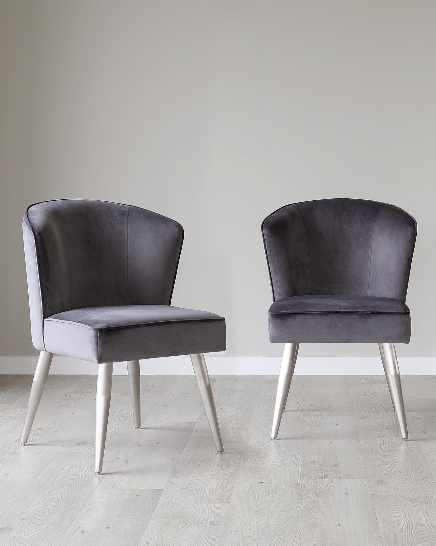 Two contemporary velvet dining chairs with high, rounded backrests and silver metal tapered legs on a light wood floor against a neutral wall background.