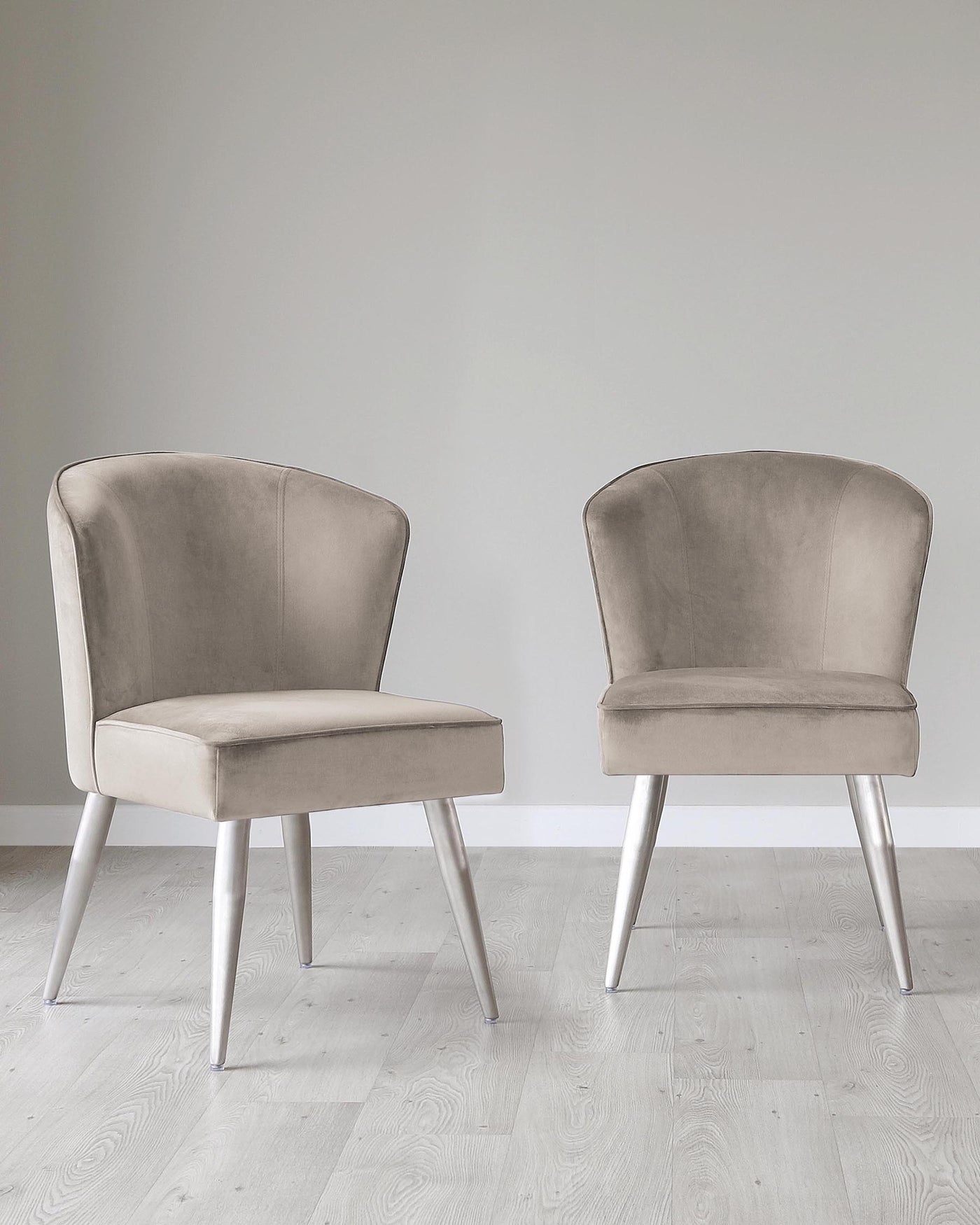 Two contemporary style dining chairs with taupe upholstery and tapered metallic silver legs on a light hardwood floor against a neutral wall.
