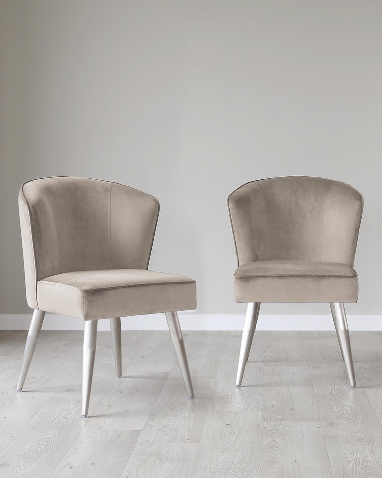 Two contemporary style dining chairs with taupe upholstery and tapered metallic silver legs on a light hardwood floor against a neutral wall.