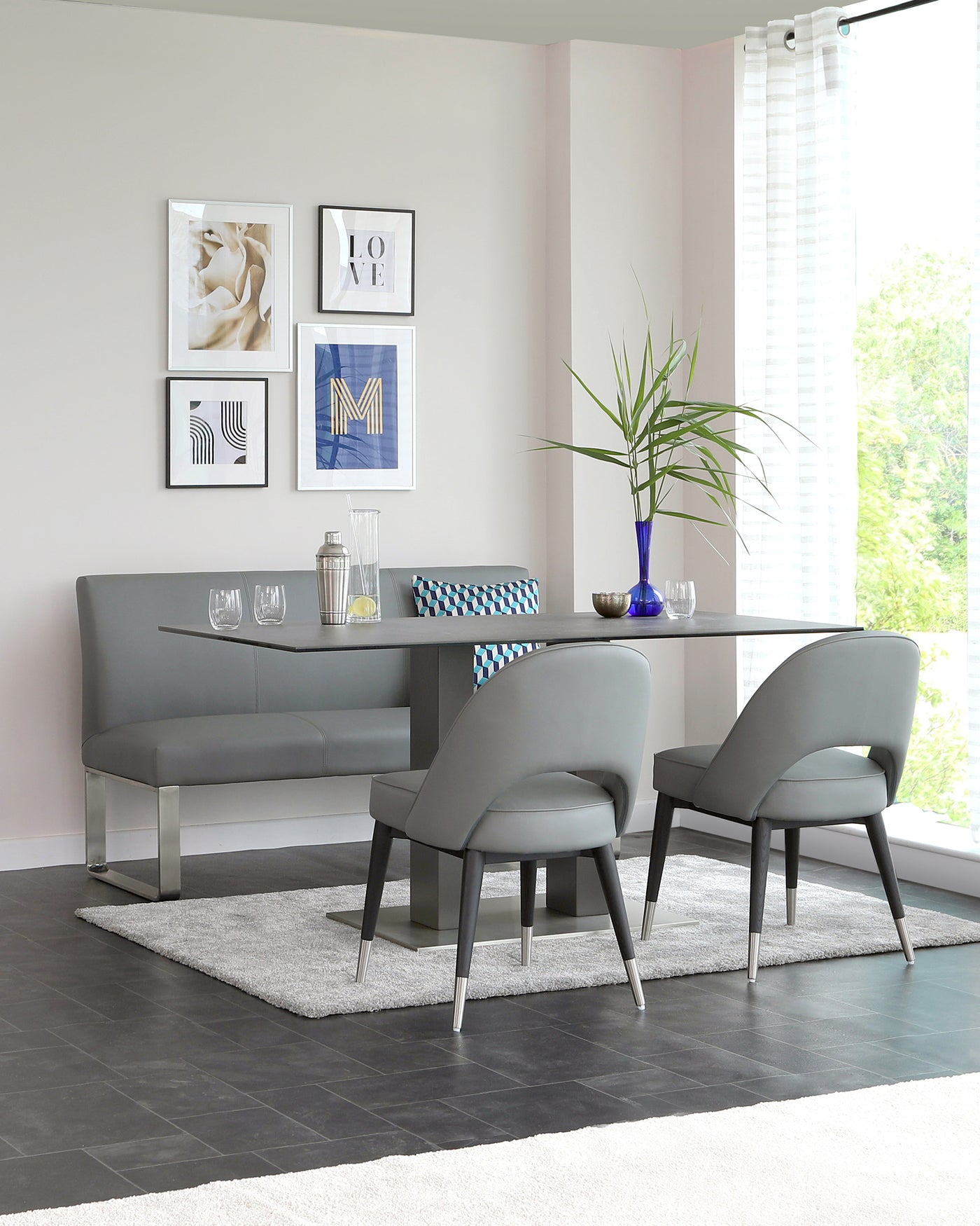 Modern dining room furniture set featuring a sleek, rectangular table with a glass top and metal legs, complemented by a long, straight-lined bench and two curvaceous chairs upholstered in a matching grey fabric, all positioned on a soft grey area rug.