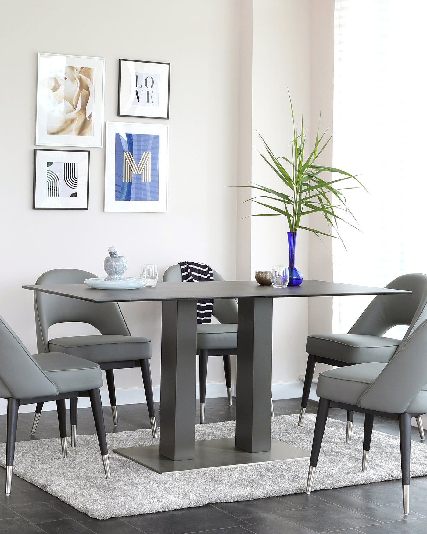 Contemporary dining room set featuring a rectangular table with a sleek, grey finish and a solid base that includes a storage shelf. The set is accompanied by four modern, upholstered dining chairs with curved backs and tapered metal legs.