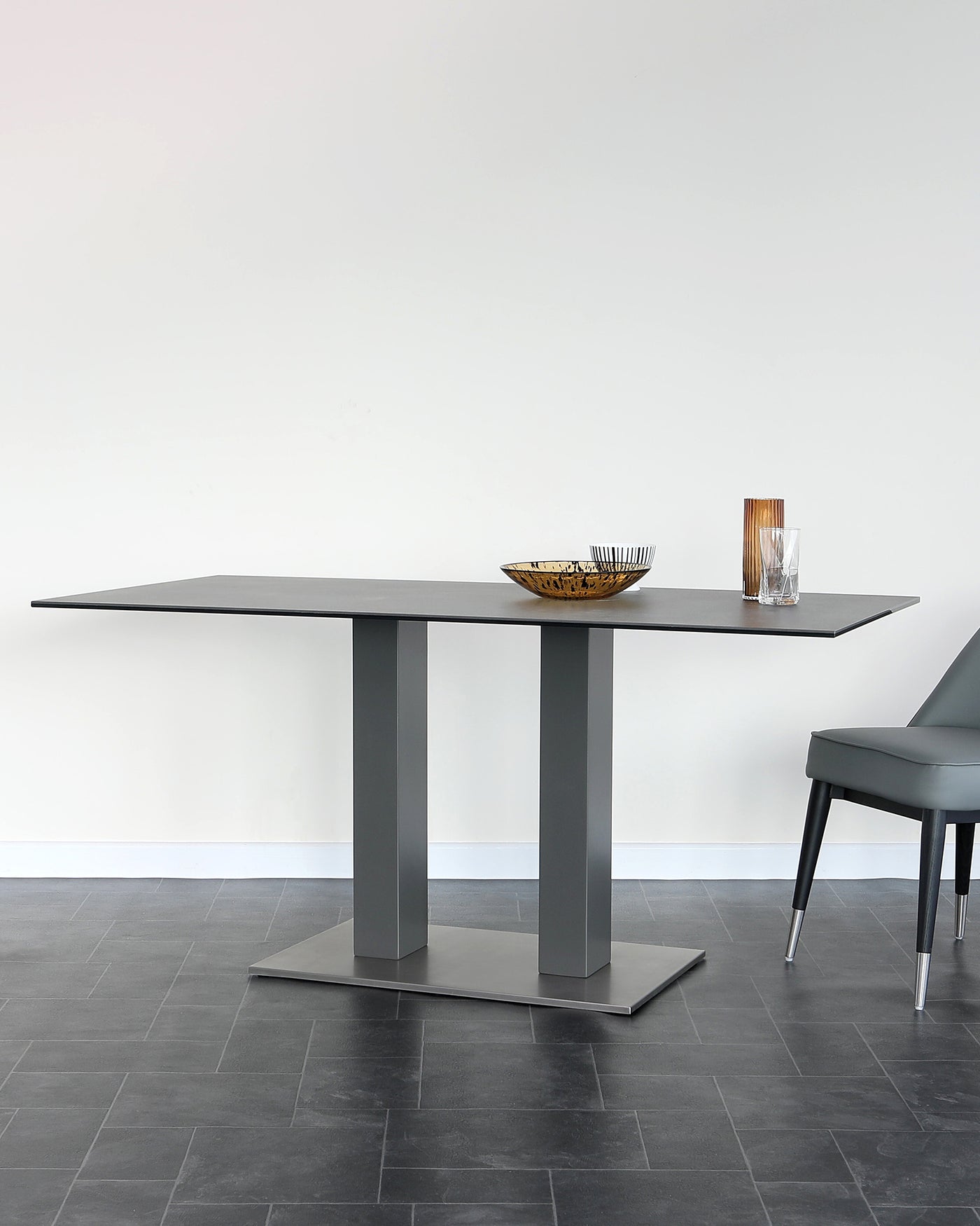 Modern minimalist dining set featuring a rectangular table with a flat, sleek surface and twin pedestal base in a matte grey finish. A single grey upholstered chair with a cantilever design and metal legs is partially visible. The table is accessorized with a decorative bowl and two drinking glasses. The set is positioned on a dark tile floor against a white wall backdrop.