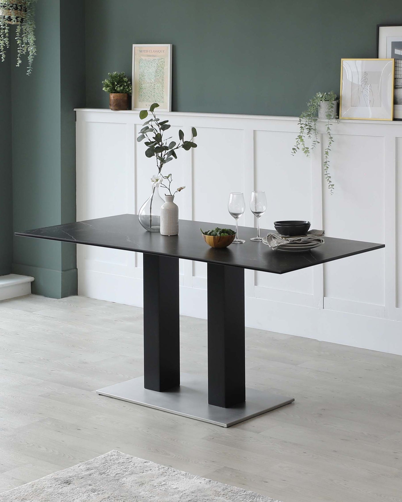 Modern rectangular dining table with a sleek black tabletop supported by two wide, vertical slab legs in a matte black finish, set upon a flat, brushed metal base. The table is staged with simple dinnerware, a textured vase holding eucalyptus branches, and paired with a neutral-toned room decor.