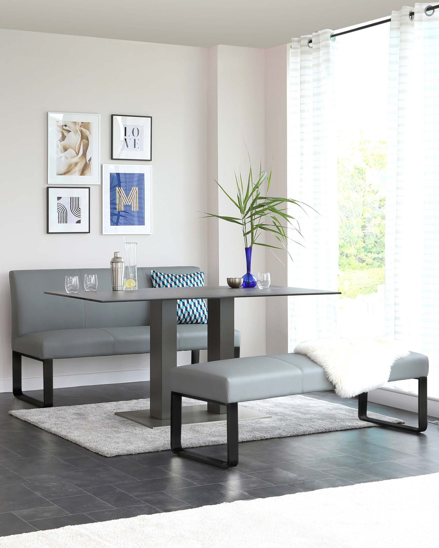 A modern dining set featuring a grey rectangular table with black metal legs, paired with a matching dining bench and two stools. The table is accessorized with decorative items including glasses, a blue vase, and a small plant. A plush grey area rug lies beneath the set.