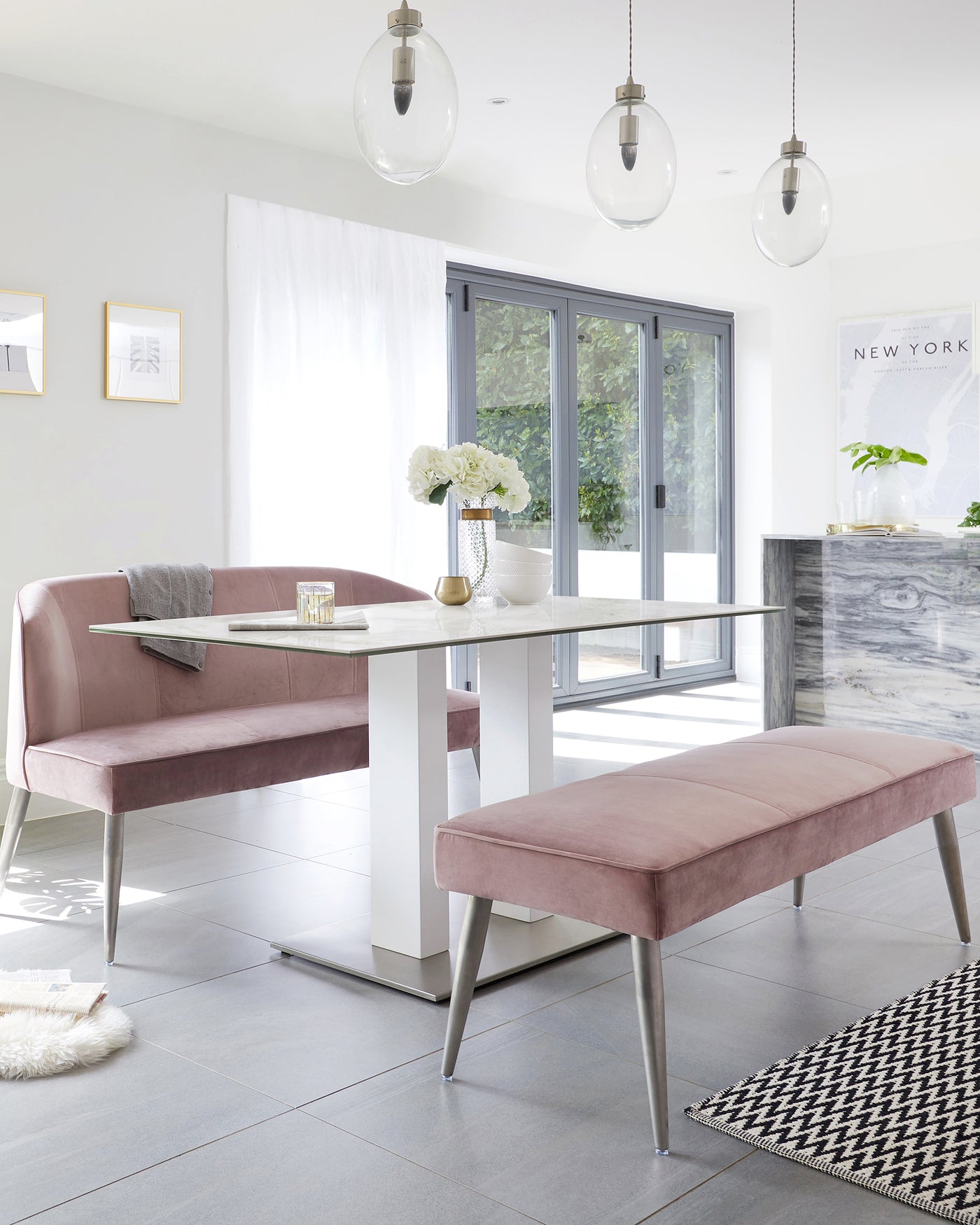 Modern dining space featuring a blush-coloured upholstered corner bench with backrest, a complementary bench without a backrest, and a rectangular white marble-topped dining table with a sleek white base and metallic silver legs.