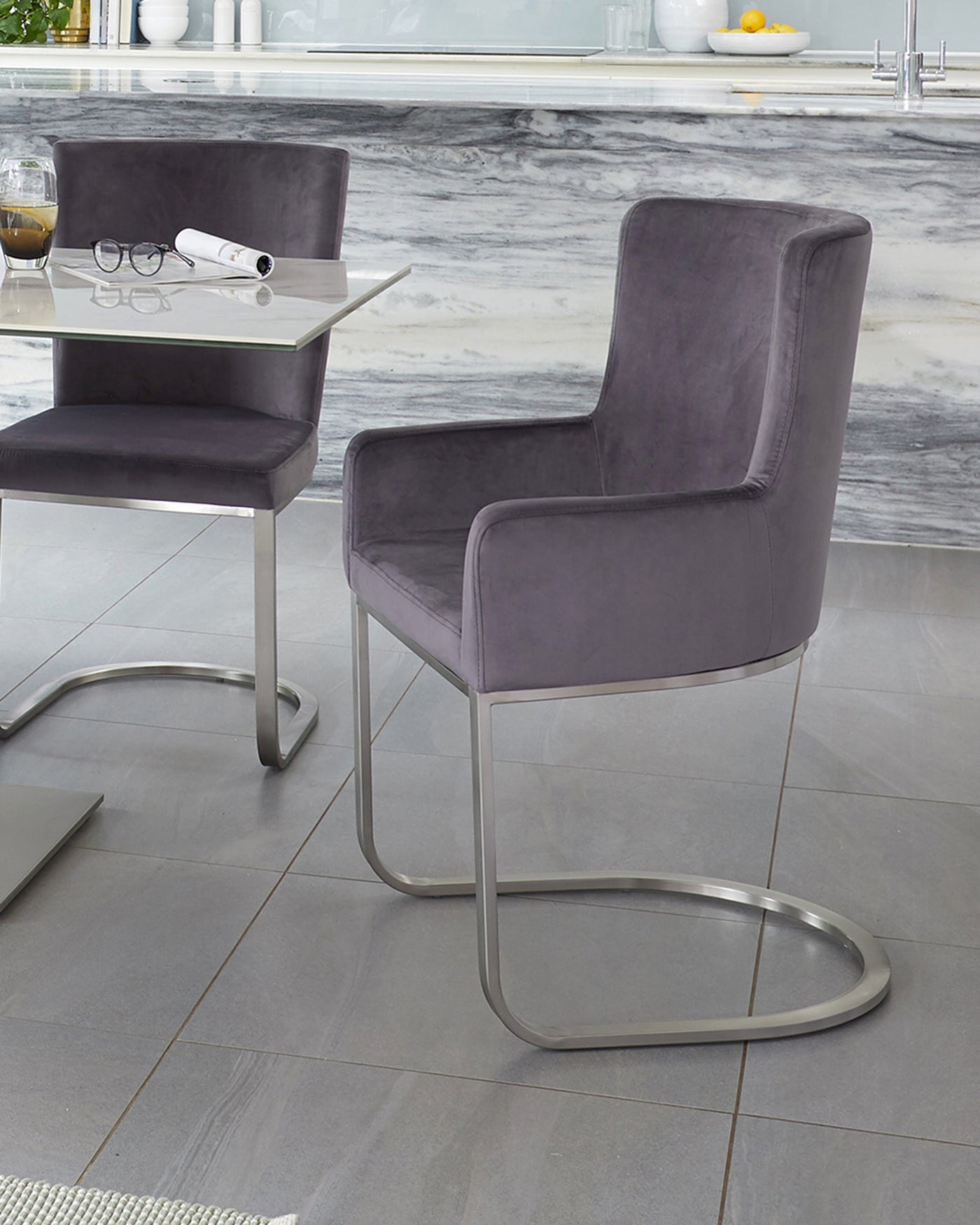 Modern furniture collection featuring a glass accent table with a chrome frame and two contemporary cantilever dining chairs upholstered in a plush grey fabric with a smooth metallic base.