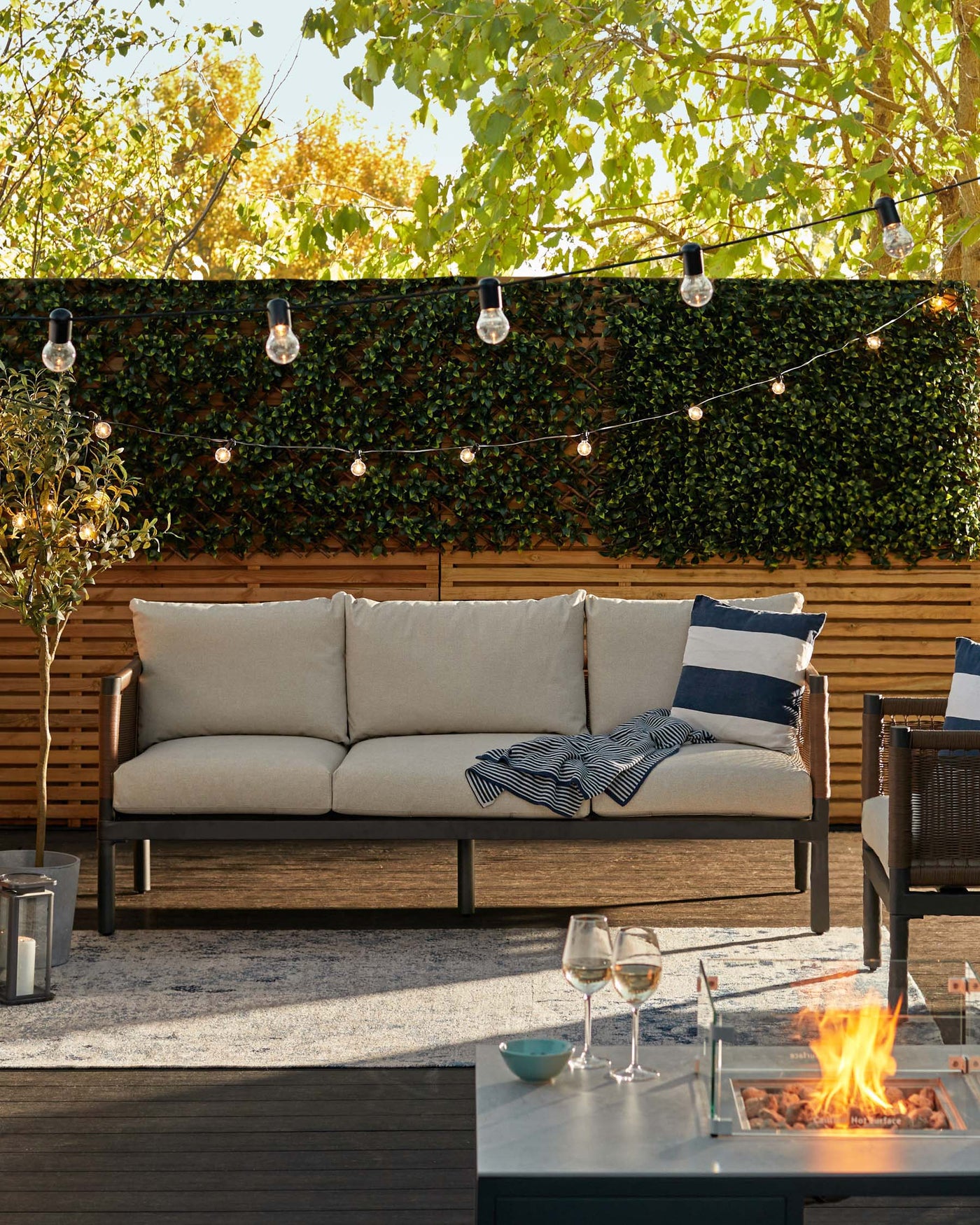 Modern outdoor furniture on a patio, including a sleek wood-frame sofa with plush beige cushions and throw pillows, a rectangular fire pit table with a glass barrier around the flame, and a matching dark grey armchair with similar cushion styling. Decorative string lights hang overhead, and the setting is complemented by a textured area rug under the sofa.