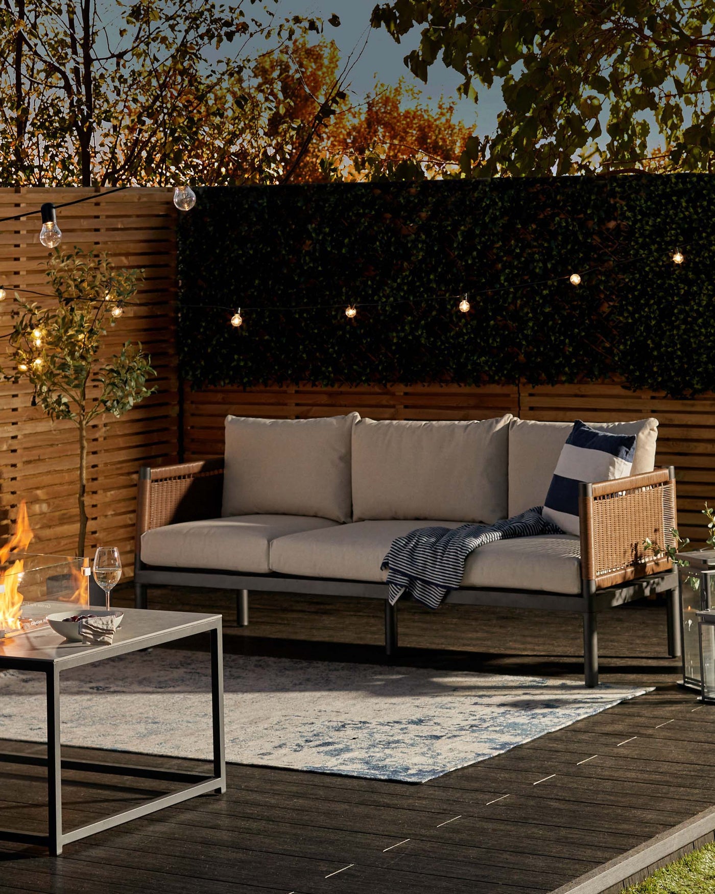 Outdoor patio setting featuring a modern sectional sofa with a metal frame and light wooden armrests, complemented by neutral-toned cushions. In front of the sofa is a sleek, square metal coffee table. The arrangement is centred on an area rug with a faded blue and cream pattern. Decorative string lights add ambiance above the cosy setup.