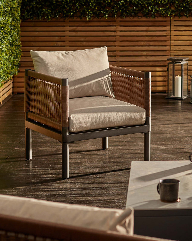 Modern outdoor armchair with a beige cushion, featuring a woven back design and metallic frame, displayed on a wooden deck with a lush hedge backdrop and decorative lantern.