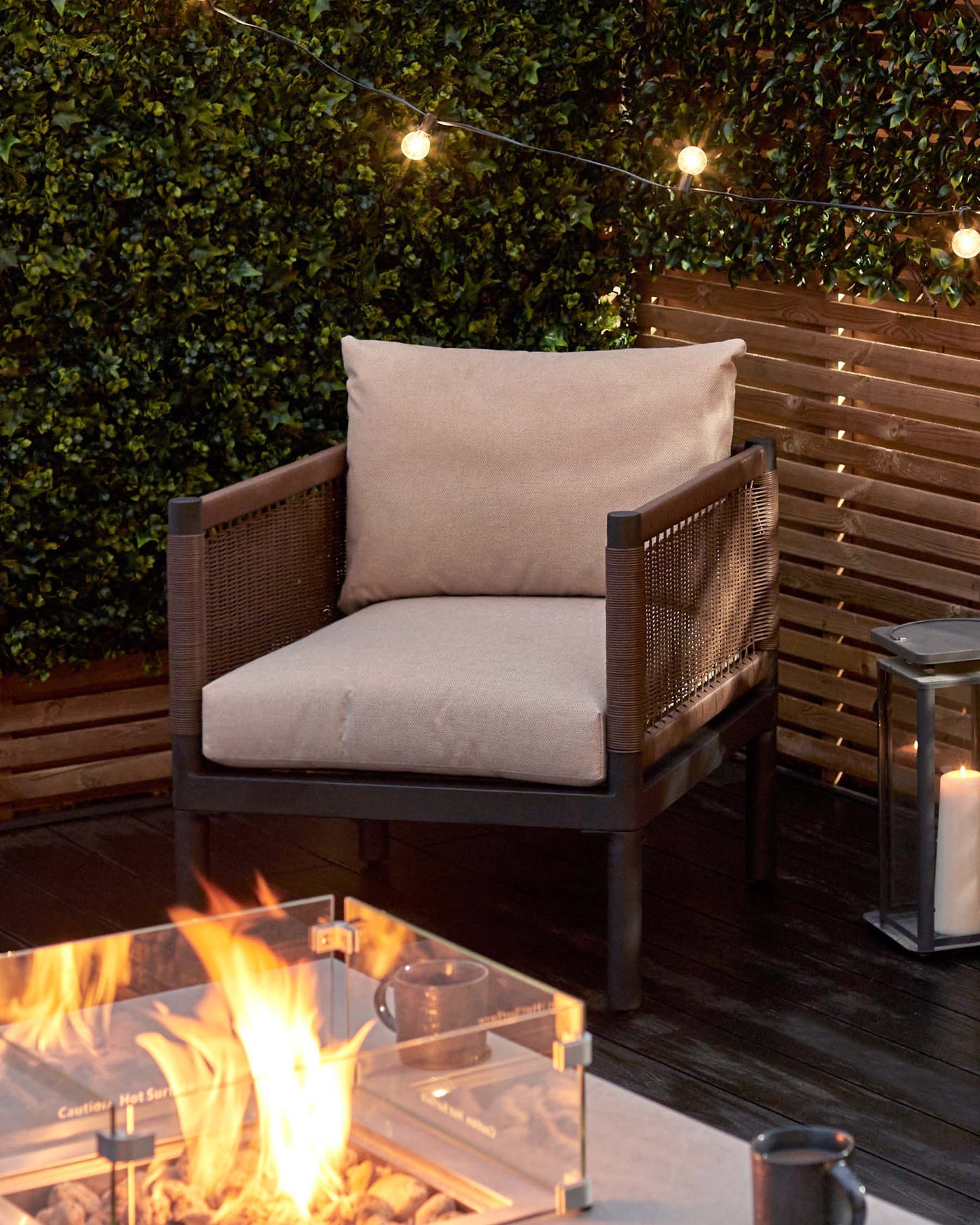Outdoor lounge chair with a modern design, featuring a sturdy dark frame with wicker accents and thick, comfortable beige cushions, set against an inviting patio backdrop with ambient string lights and a glowing fire pit table.