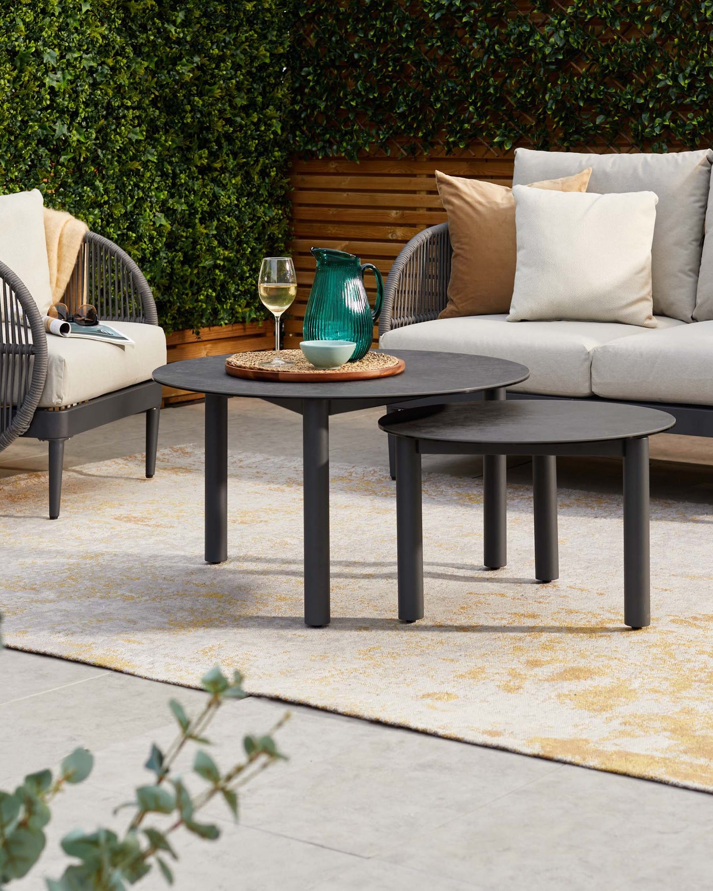 Outdoor furniture set featuring a modern grey two-seater sofa with plush cushions and a pair of round, black nesting coffee tables on a neutral-toned area rug.