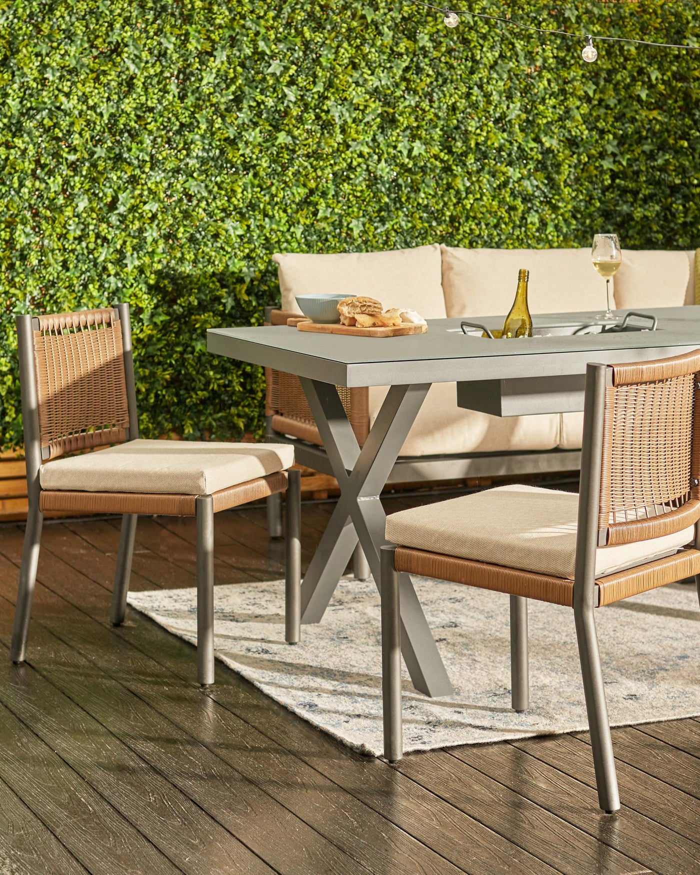 Outdoor patio furniture set featuring a modern grey dining table with a unique X-shaped base and two beige upholstered dining chairs with grey metal frames and wicker panel accents, arranged on a faded print area rug over wooden decking, against a backdrop of a lush green hedge. A sofa with off-white cushions is partially visible.