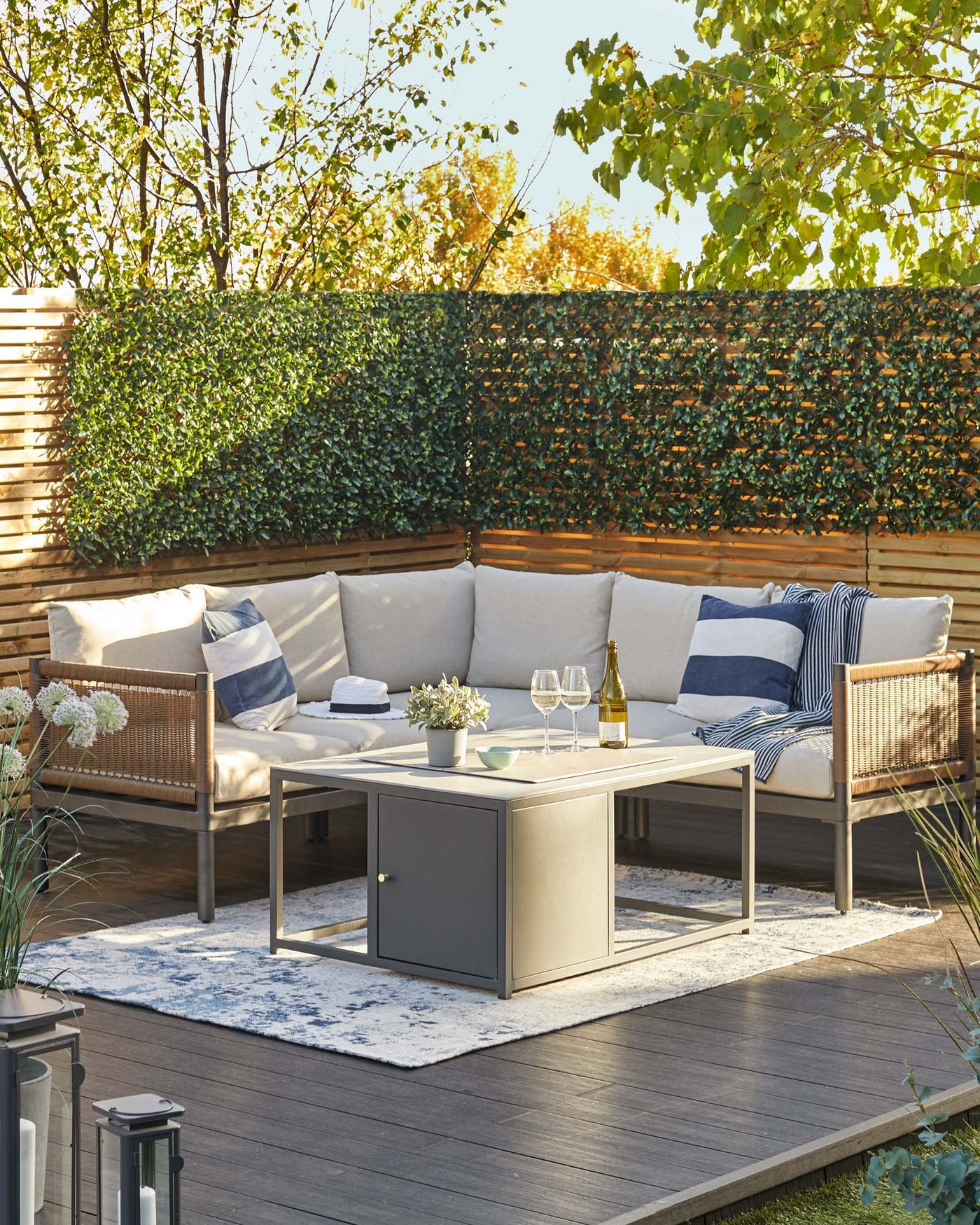 Outdoor furniture set on a wooden deck, featuring a modular corner sofa with light beige cushions, and a modern, rectangular coffee table with a grey finish and storage compartment. Accessorized with striped and solid throw pillows, white flowers, and glassware.