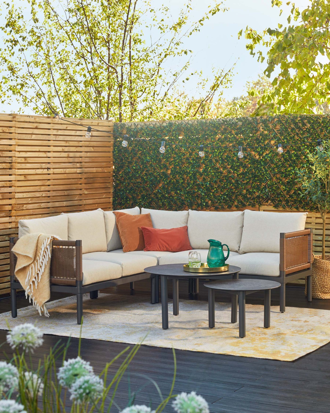 Outdoor patio furniture set featuring a sectional corner sofa with beige cushions and accompanied by a single matching armchair. A central coffee table accompanied by two smaller nesting tables, all in a matching dark finish, sits atop a light, patterned area rug. The set is adorned with throw pillows in neutral and reddish tones, and a cosy blanket is draped over the armchair.