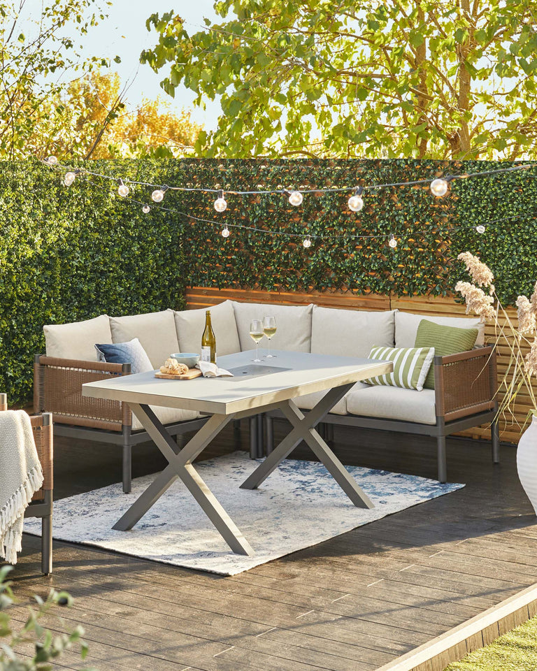 Outdoor furniture set featuring a modern, rectangular dining table with a light grey surface and X-shaped metallic legs, accompanied by a plush corner sectional sofa in a natural wicker finish with cream cushions and accent pillows in various patterns. A textured light blue and white area rug lies beneath the table, all arranged on a wooden deck with ambient string lights overhead.