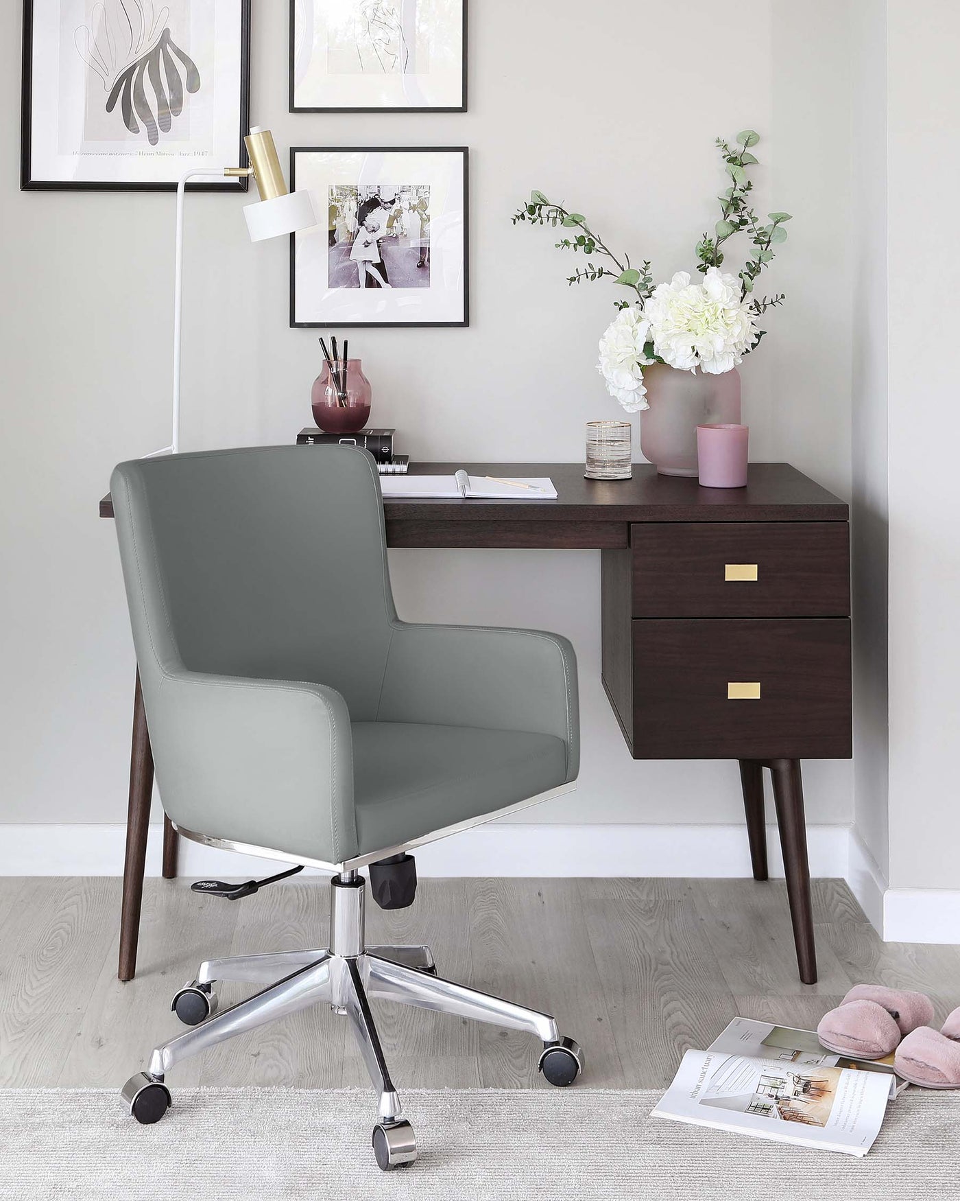 A modern home office setup featuring a sleek, mid-century inspired dark wood desk with an open work surface, one drawer, and a storage compartment with brass handle accents, paired with a comfortable, grey upholstered swivel chair with a high backrest and mounted on a five-point chrome base with caster wheels. A light area rug sits beneath the furniture ensemble.