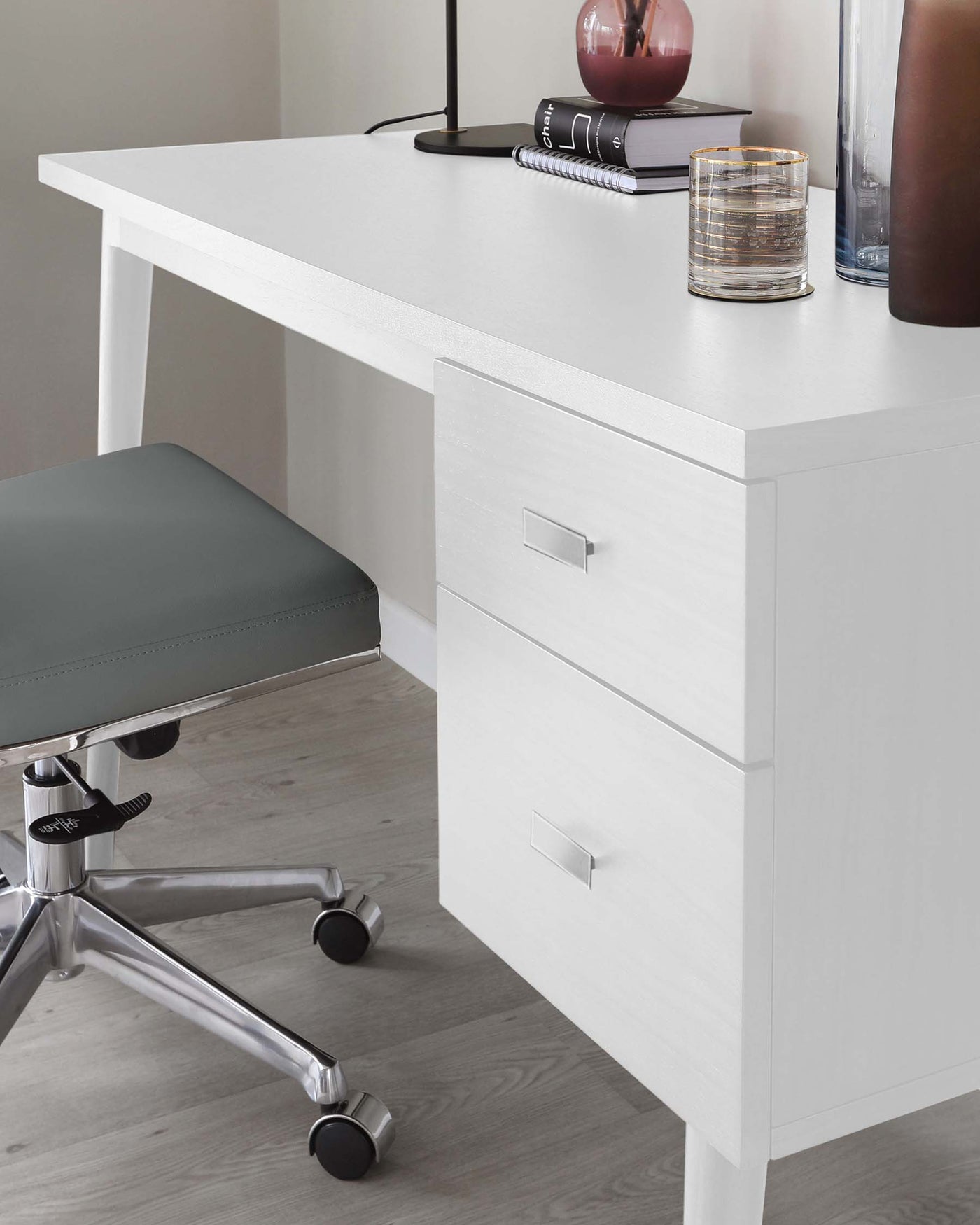 A modern white desk with clean lines, featuring an uncluttered tabletop and two drawers with simple rectangular silver handles. Paired with the desk is a grey upholstered office chair with a metallic base and caster wheels for mobility.