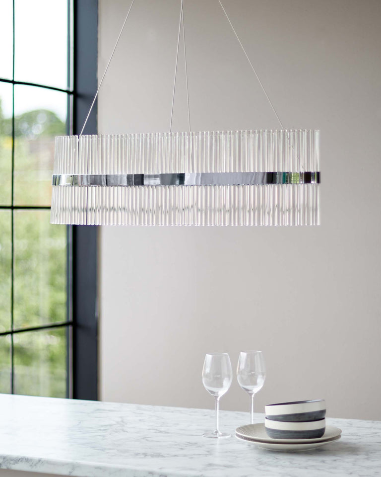 Elegant modern chandelier with vertical clear glass rods, suspended over a sleek white marble countertop, complemented by two stemware glasses and neatly stacked round plates with grey accents.