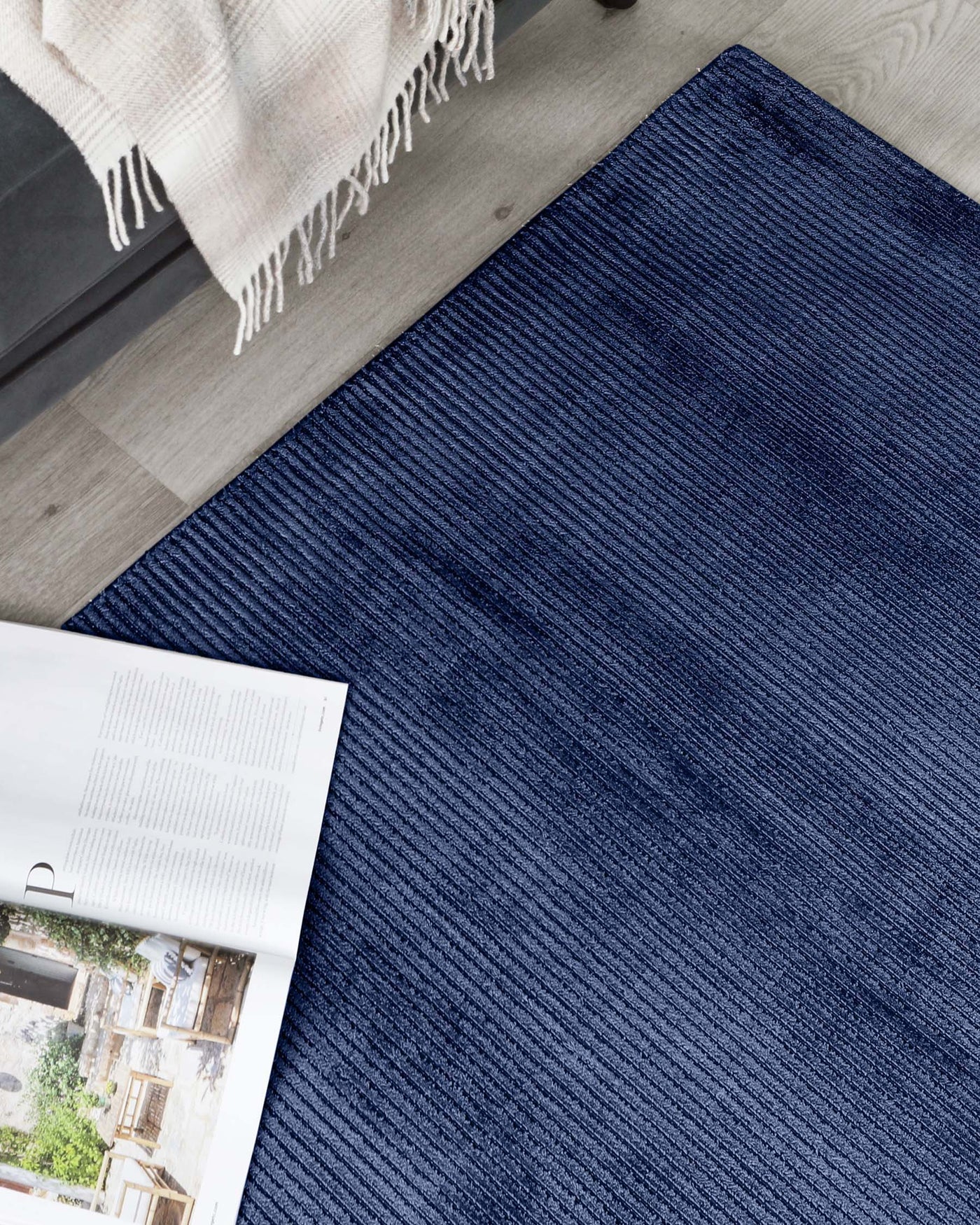 Contemporary deep blue textured area rug with a subtle linear pattern, placed on a hardwood floor partially under a light grey upholstered bench with a beige fringed throw blanket casually draped over the corner, next to an open magazine.