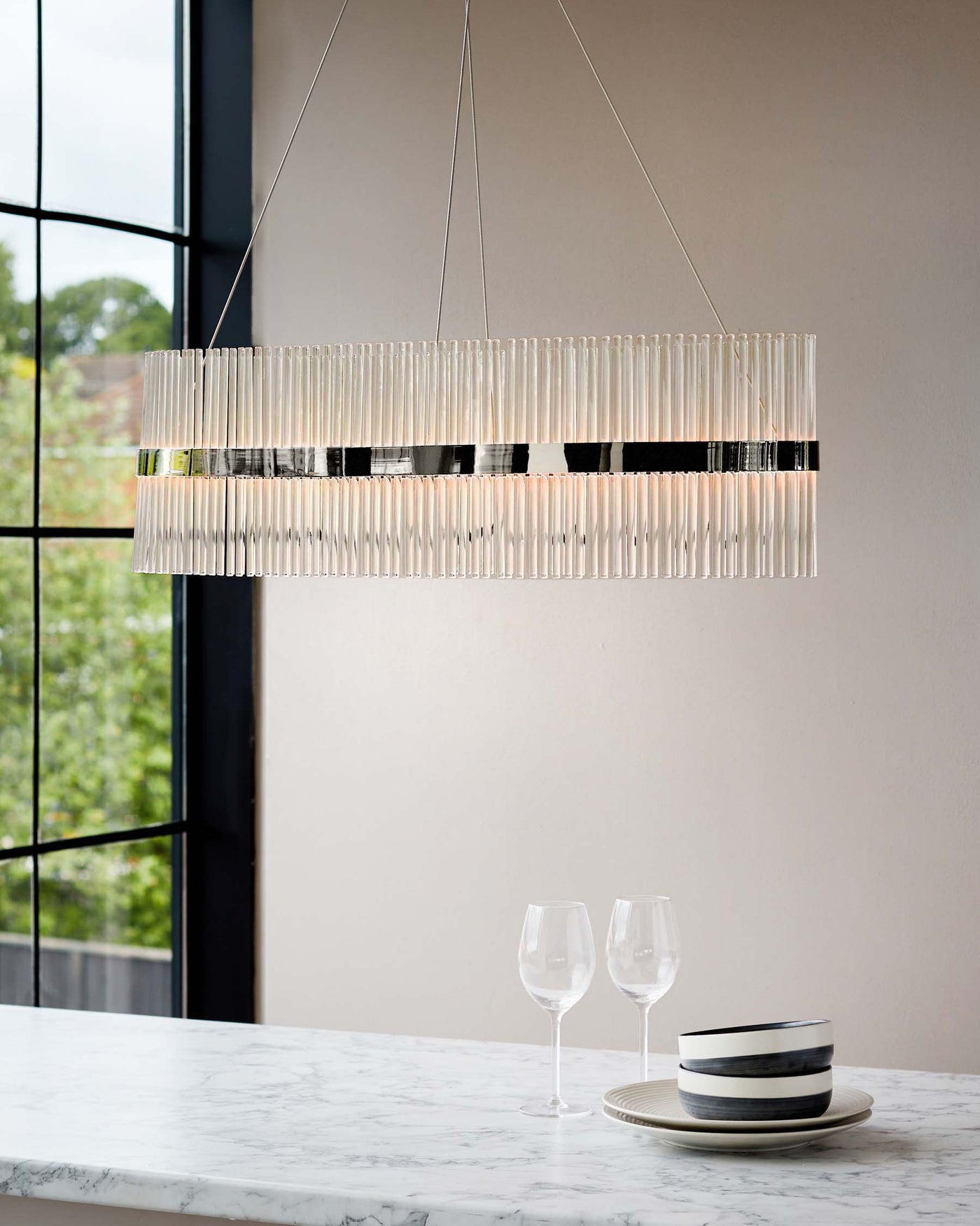 Elegant rectangular chandelier with vertical clear glass rods hanging above a sleek marble countertop, accented by two stemless wine glasses and a stack of modern two-tone plates on a round woven placemat.