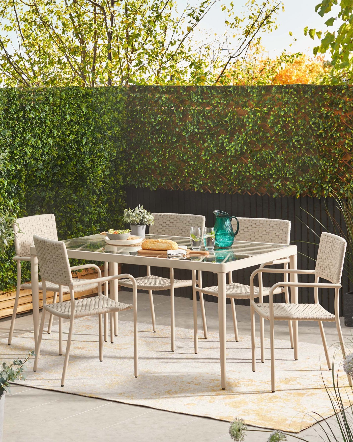 Modern outdoor dining furniture set consisting of a rectangle glass-topped table with beige metal frame and four matching armchairs with woven seat and backrest in a light, neutral tone. The set is arranged on an outdoor patio with a natural backdrop.