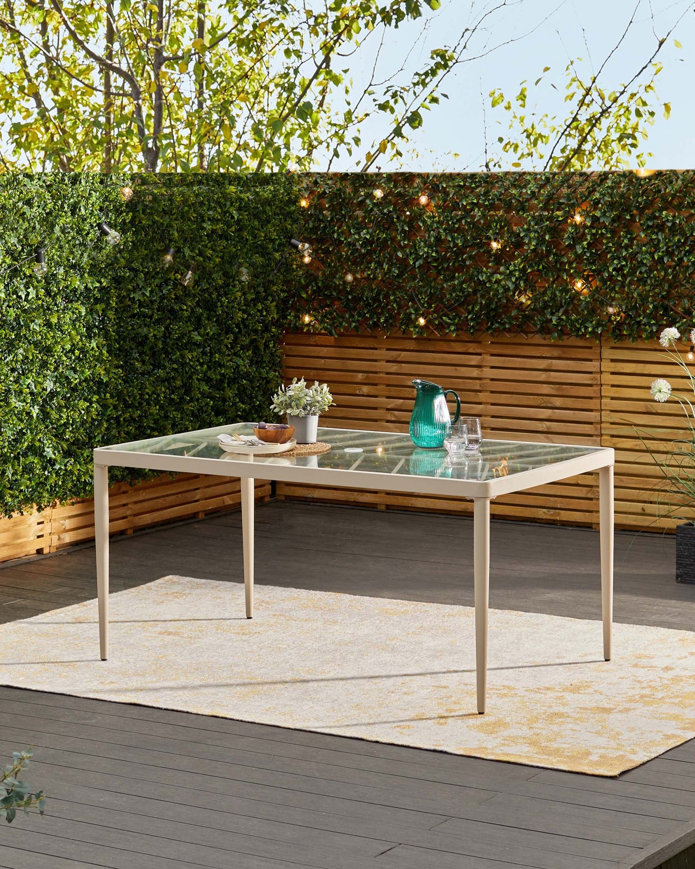 Modern outdoor rectangular dining table with a translucent glass top and beige metal frame, displayed on a cream and yellow patterned outdoor area rug.