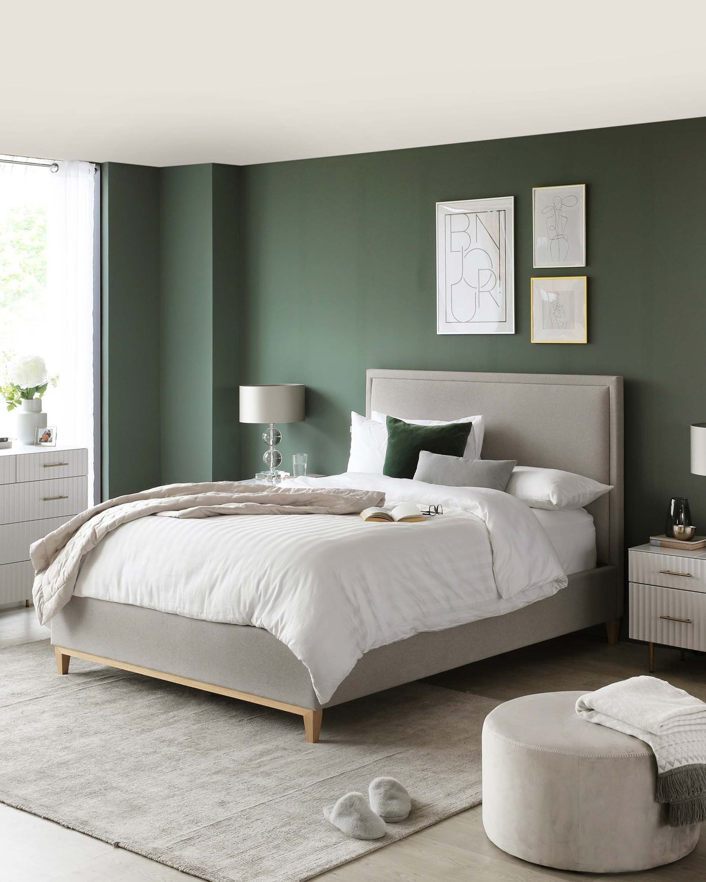 Modern bedroom with a king-sized bed upholstered in a light grey fabric featuring a simple headboard, complemented by matching light grey nightstands with white drawers, accented by sleek metal pulls. A small, round, light grey ottoman is positioned at the foot of the bed on top of a large area rug that adds a layer of texture to the room.