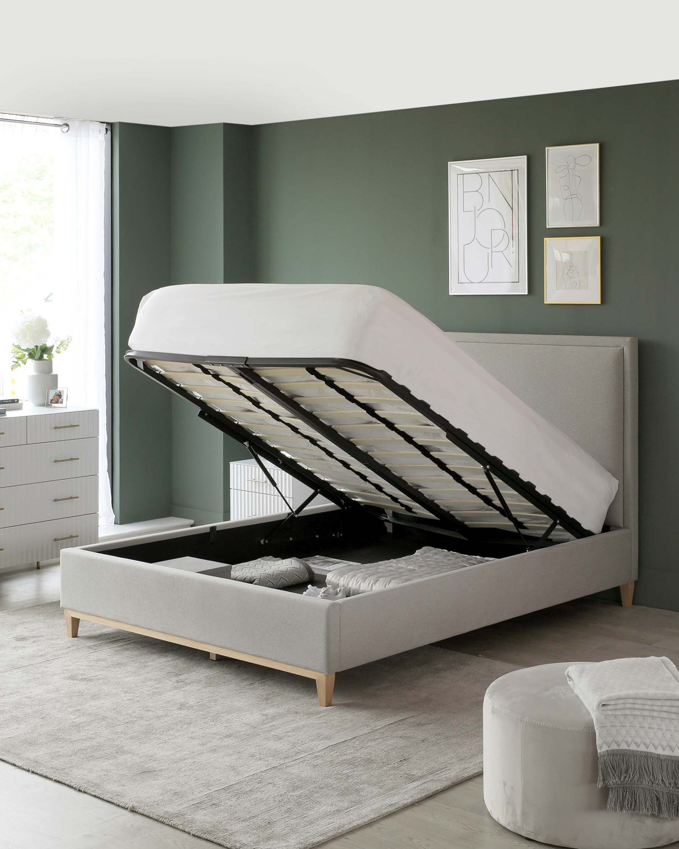 Contemporary light grey fabric upholstered storage bed with a lifted mattress revealing spacious under-bed storage, complemented by a small white bedside chest of drawers topped with a vase of flowers and a simple round, grey ottoman. The bed is accented with light wooden legs and framed art above, within a minimalist bedroom setting.
