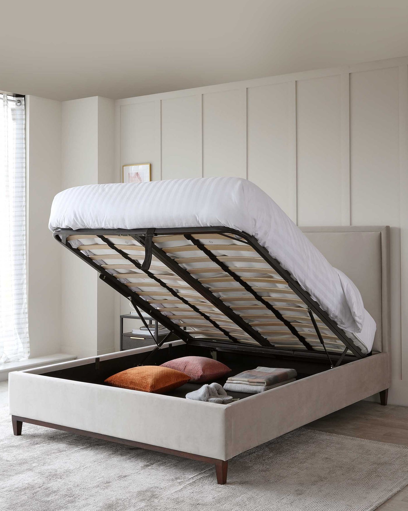 Elegant light grey upholstered storage bed with a lifted mattress base revealing an organized under-bed storage compartment containing pillows, blankets, and other linens.