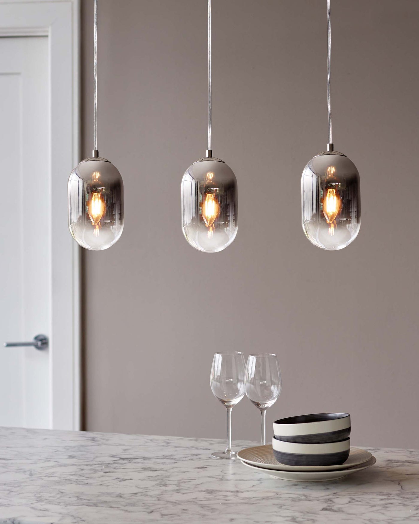 Three modern pendant lights with a sleek glass design hanging above a marble countertop displaying two wine glasses and a set of stylish striped ceramic bowls and plates.
