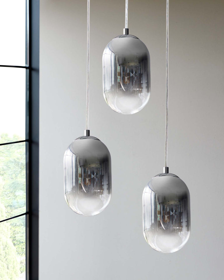 Three contemporary pendant lights featuring elongated, oval-shaped glass shades with a subtle metallic finish, suspended by slender cables against a backdrop of large windows with a view of greenery.
