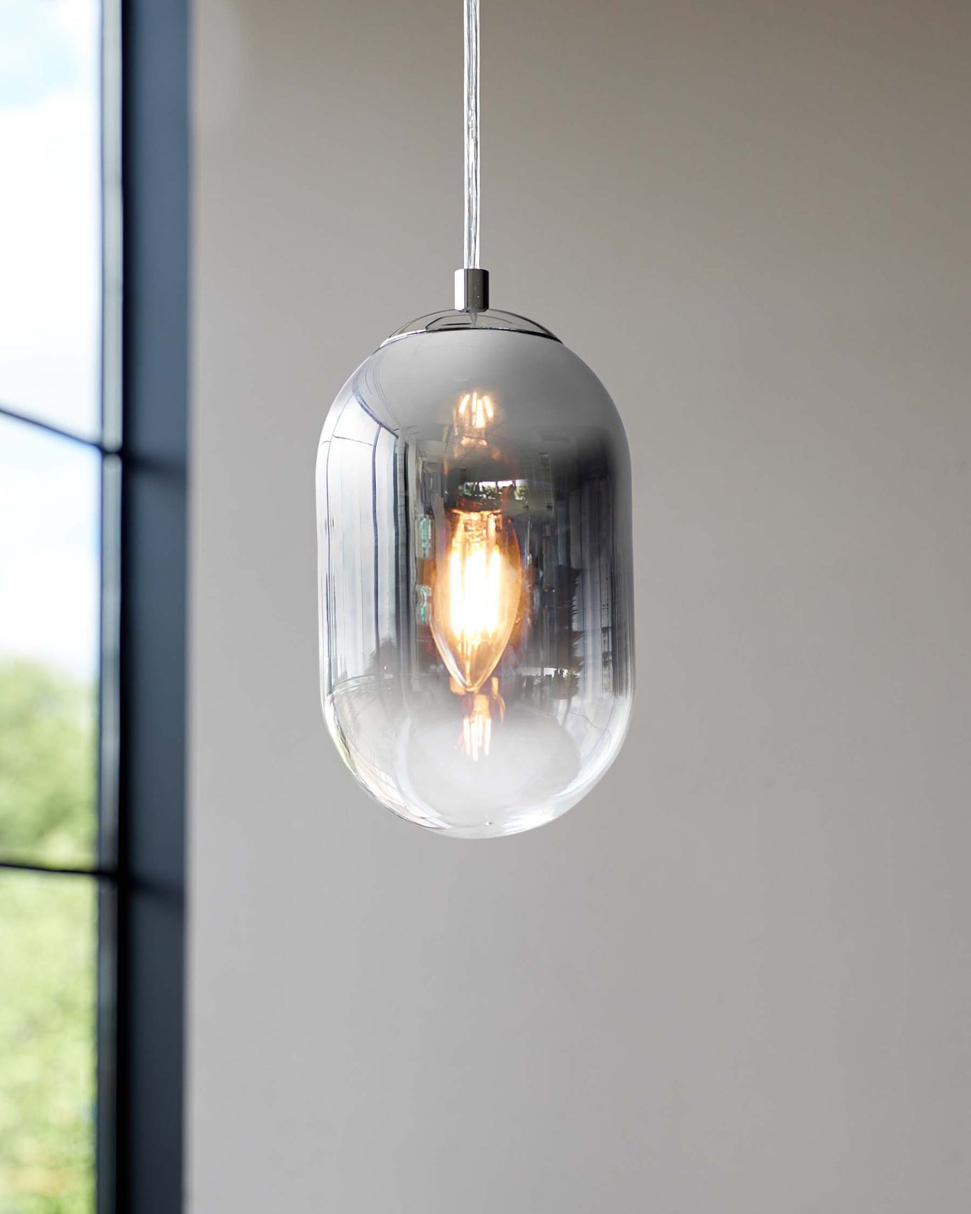 Suspended pendant light with a sleek chrome finish featuring an elongated clear glass shade encasing a visible filament bulb, set against a soft grey background near a window.