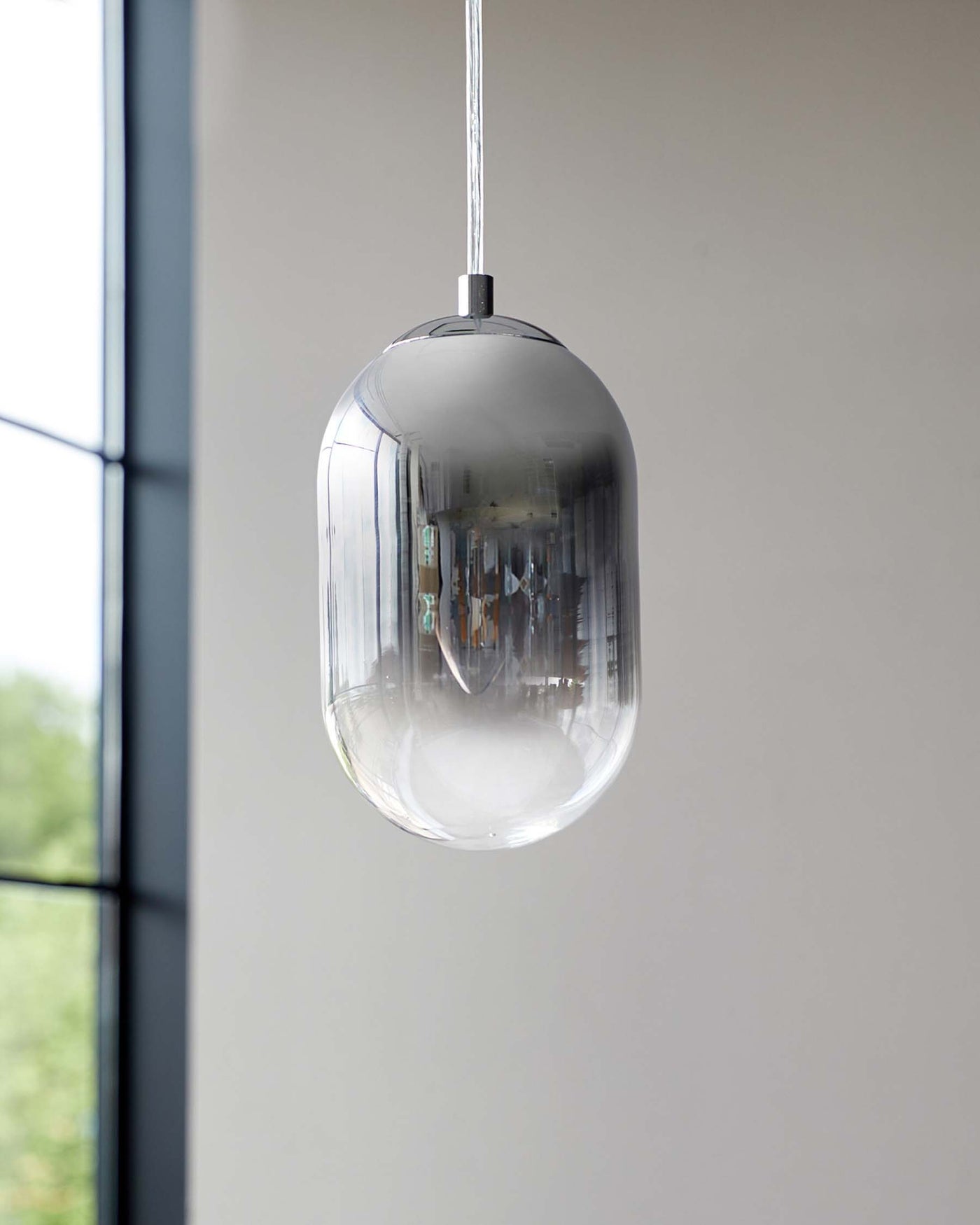 Modern clear glass pendant light with sleek metal fixtures, suspended by a wire against a soft white backdrop near a window with a view of greenery.