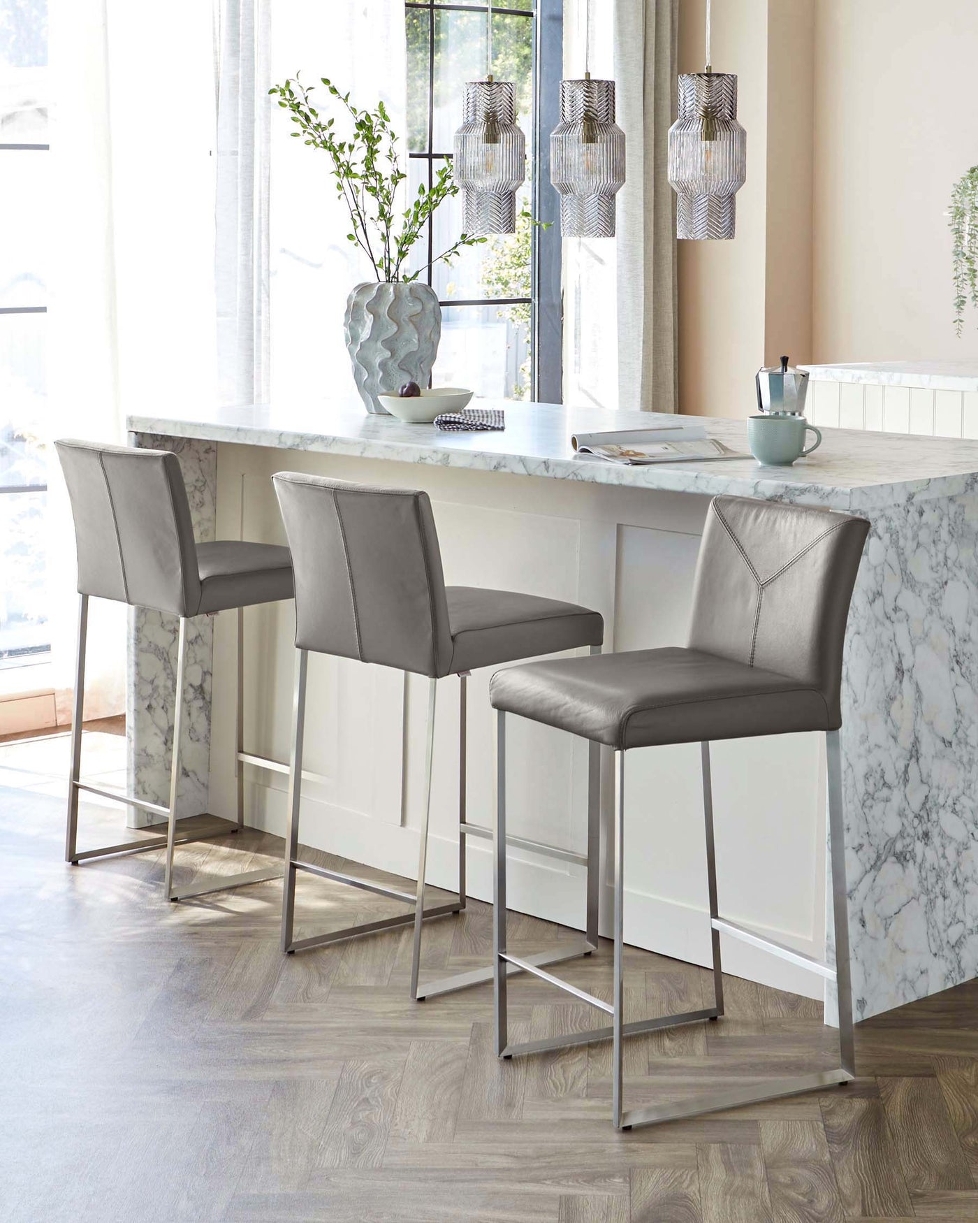 Two contemporary upholstered bar stools with sleek, silver-finished metal legs positioned at a high marble countertop. The stools feature a mid-back design with clean lines and are upholstered in a light grey fabric, complementing the bright, modern kitchen setting.