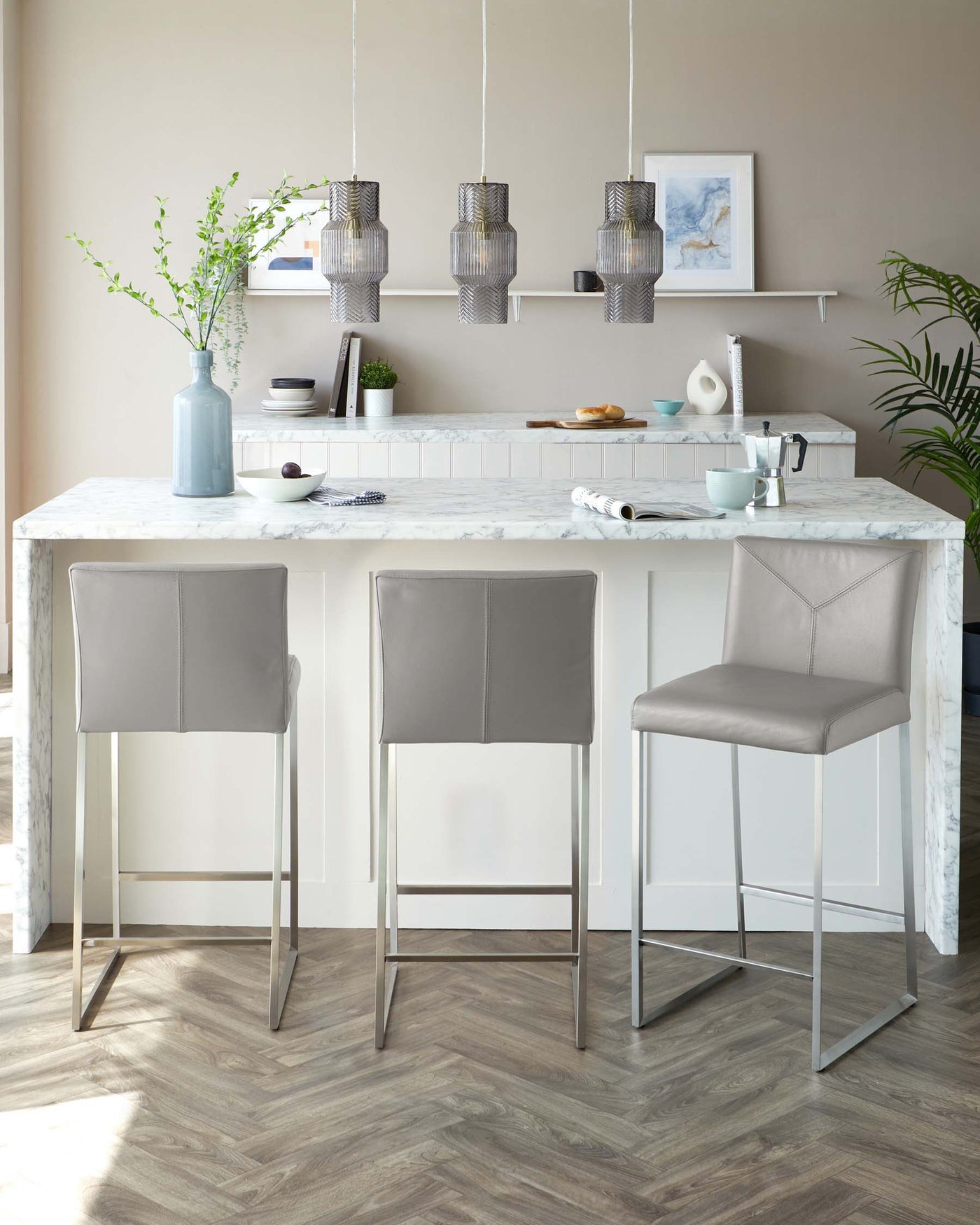 Three contemporary grey upholstered bar stools with slender metallic legs lined up at a white marble-topped kitchen island.