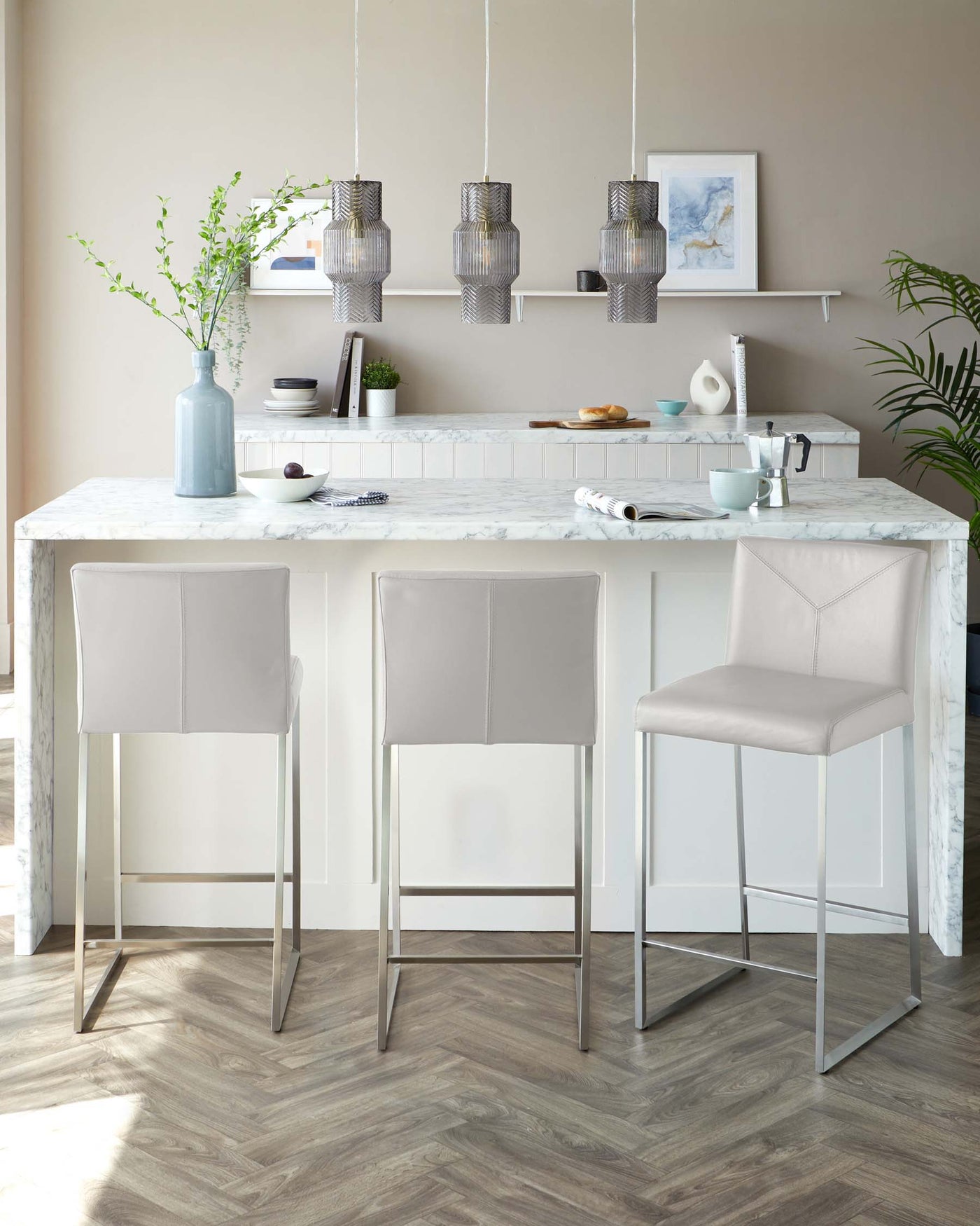 Three modern light grey upholstered bar stools with sleek, silver metal legs, positioned at a white marble-topped kitchen island.