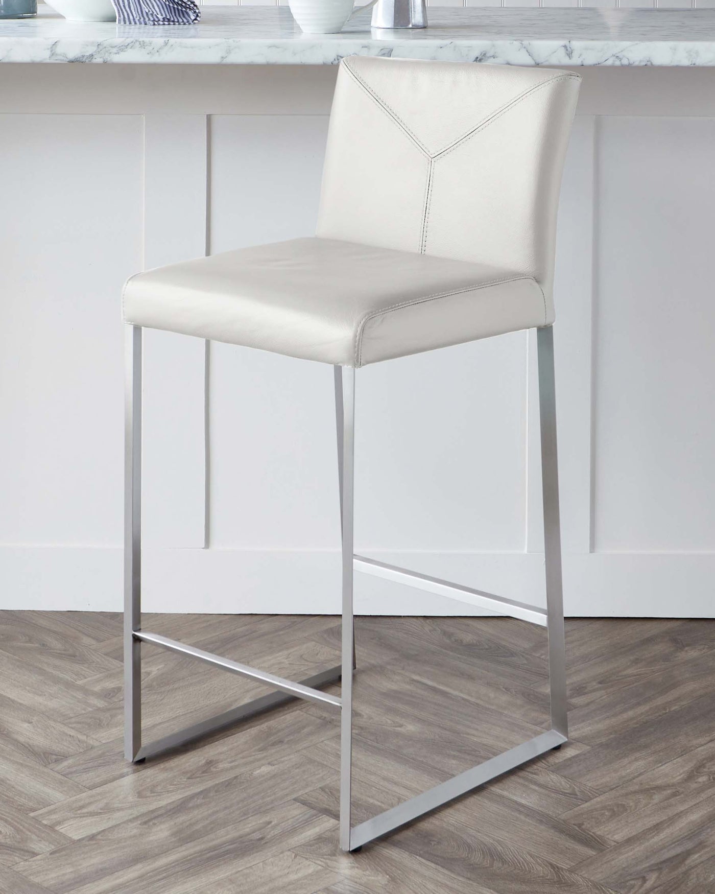 Modern bar stool with a light beige, leather-like upholstered seat and backrest, featuring a geometric stitch pattern on the back, complemented by a sleek chrome-finished metal frame with a simple footrest.