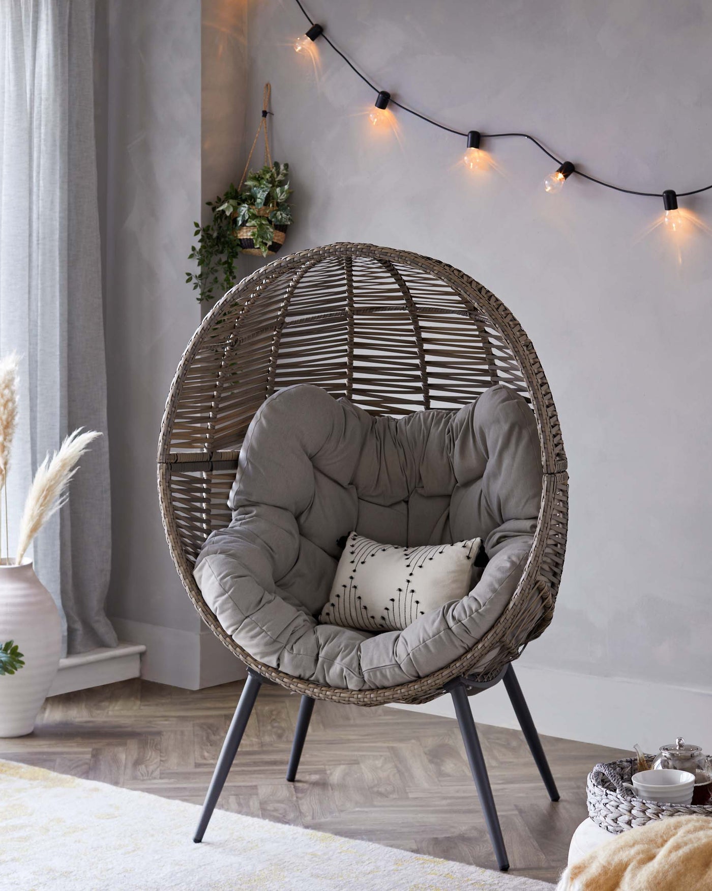A contemporary egg-shaped wicker chair with a plush grey cushion, standing on four sleek black metal legs. A small rectangular patterned pillow adds an accent to the cosy nest-like seat, which offers a stylish and inviting place to relax.