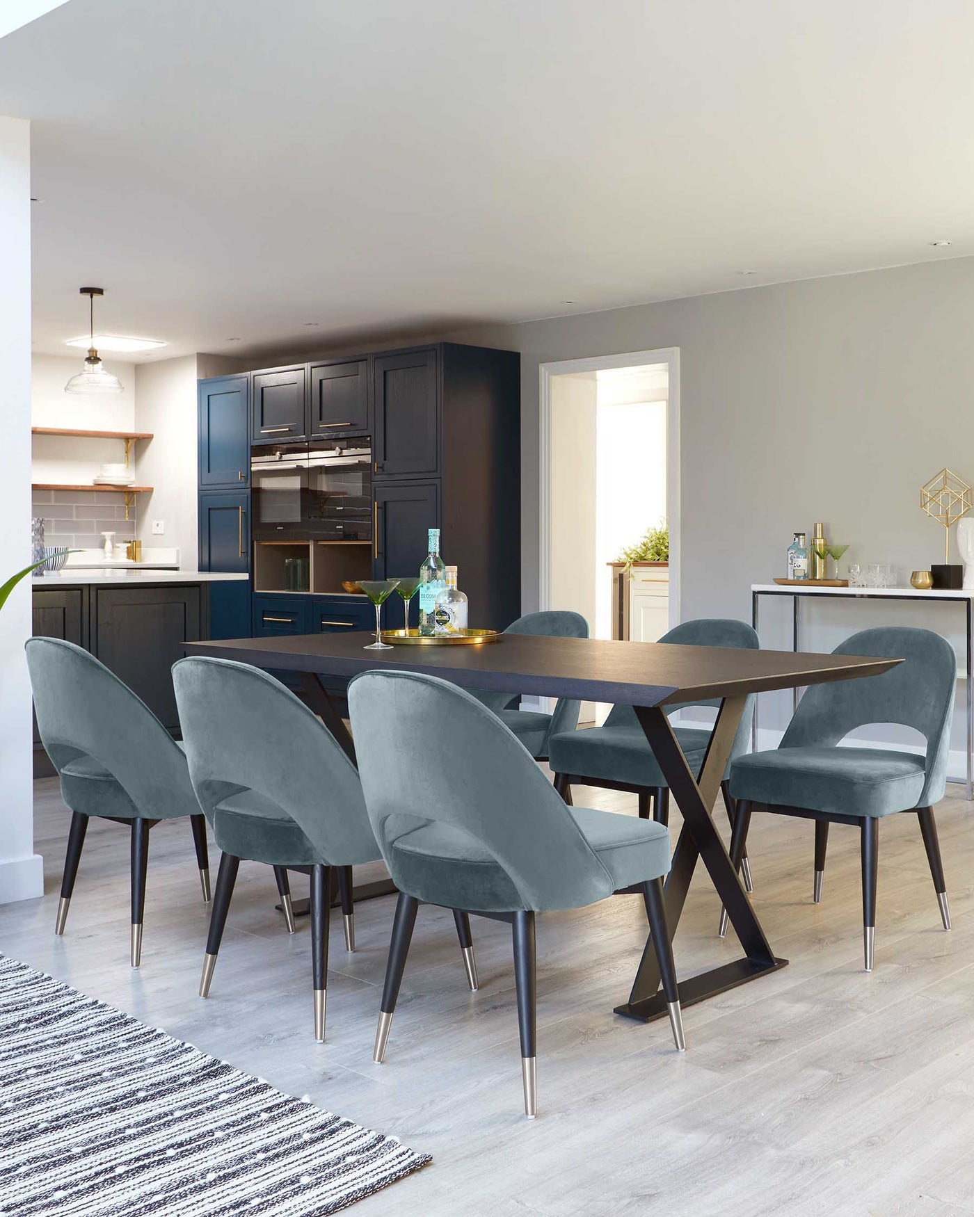 Modern dining room featuring a dark wood rectangular table with a unique X-shaped base design and metal tips, surrounded by six plush velvet dining chairs in a soft grey colour with sleek black wooden legs tipped with metallic accents. The room is styled with a light hardwood floor and a neutral colour scheme.