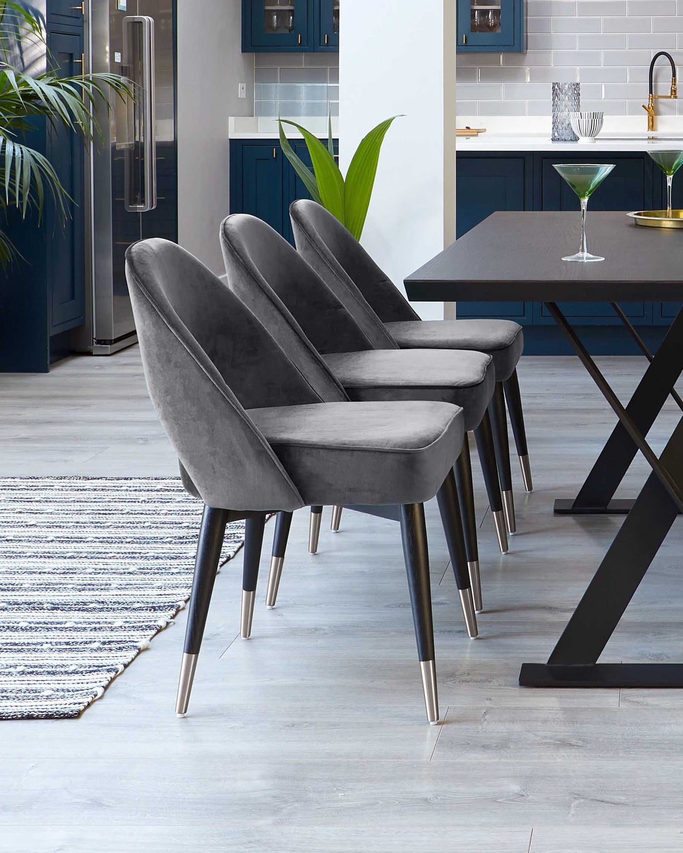 A set of four modern dining chairs with dark grey velvet upholstery and angled metal legs with gold-toned tips, paired with a minimalist dark wooden dining table with a rectangular top and sturdy black metal legs, displayed on a light wooden floor with a textured grey and white area rug. The setting is complemented by a contemporary kitchen with blue cabinets in the background.