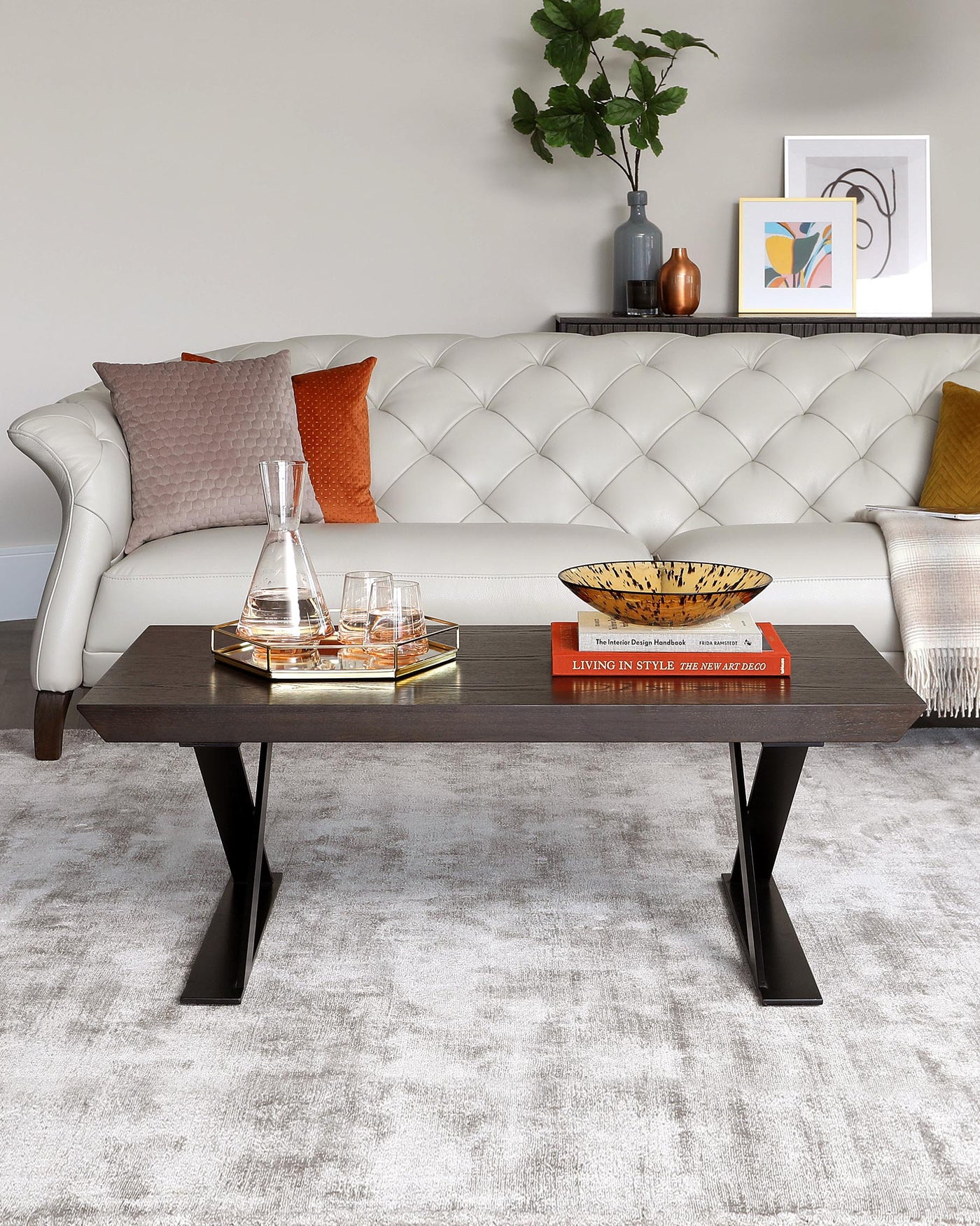 Elegant modern living room featuring a button-tufted white leather Chesterfield sofa with grey and orange throw pillows, accompanied by a dark wood coffee table with bold, angular black metal legs. The table displays a decorative tray with whisky glasses and a decanter, flanked by colourful hardcover books.