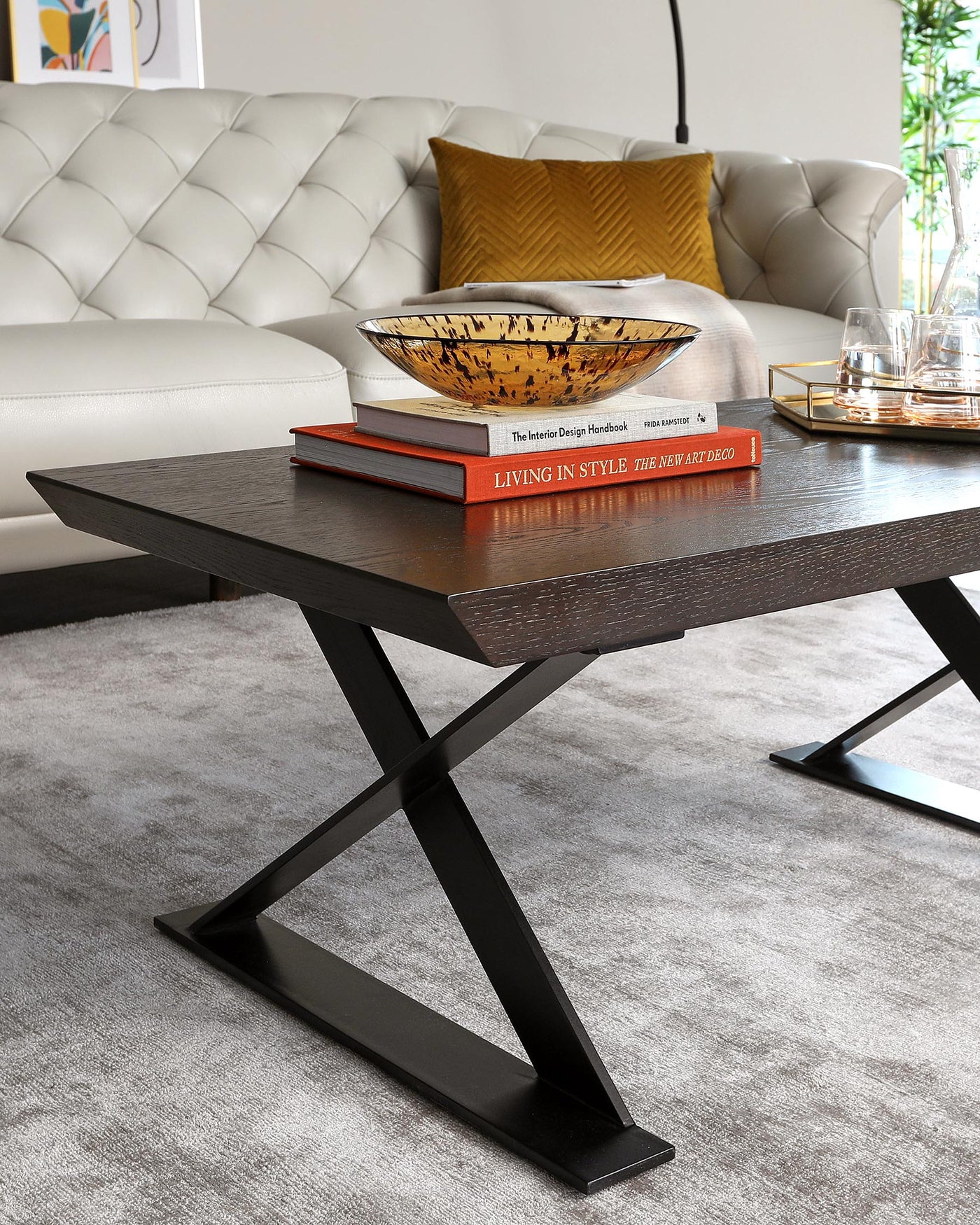 Modern coffee table with dark wood finish and distinctive geometric black metal legs, placed on a plush area rug in a well-appointed living room, accessorized with decorative books and an ornamental bowl.